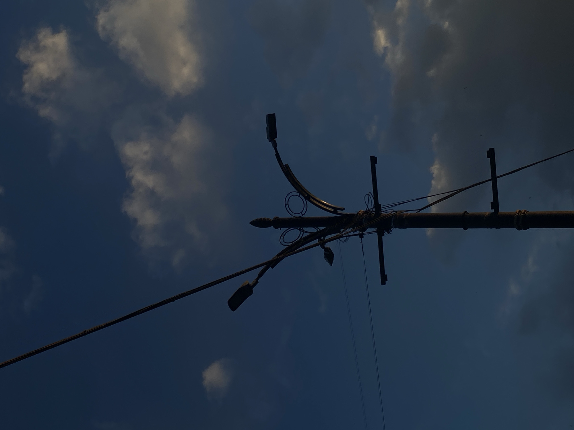 A low-angle shot depicts a silhouetted utility pole extending horizontally across the frame. The pole supports various wires and two streetlights, one curving upward and the other downward. The background is a blue sky with white clouds.