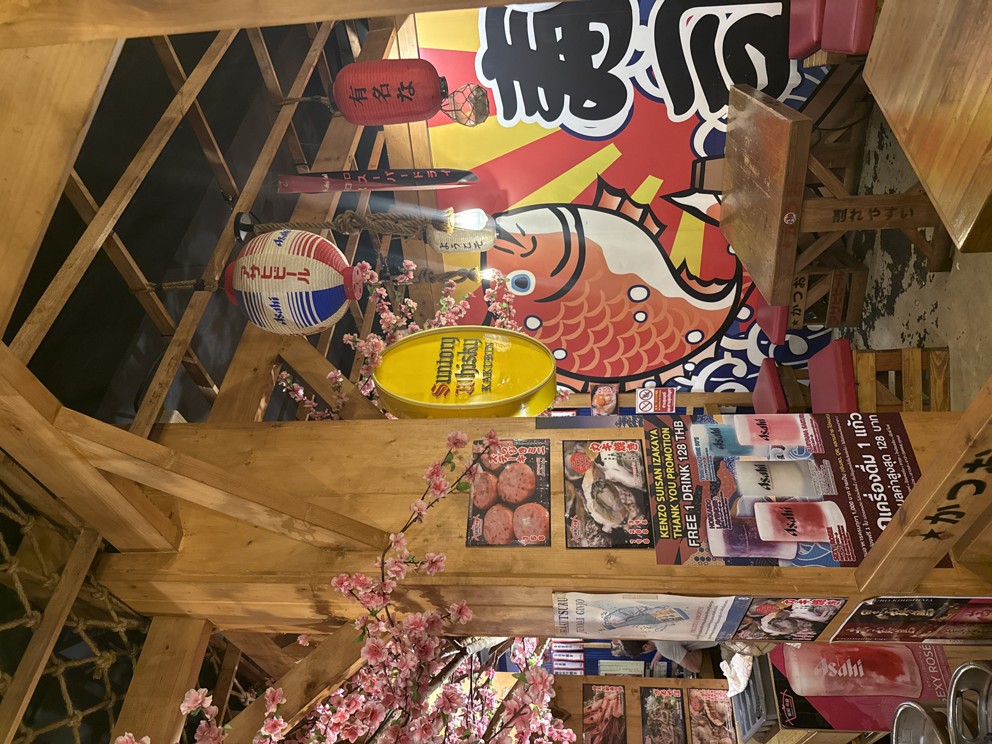 The image shows the interior of a Japanese-style izakaya or restaurant in Bangkok, Thailand, with a focus on the ceiling area. The ceiling is decorated with a variety of Japanese items including red and white paper lanterns, a large poster with Japanese text and a drawing of a fish, a yellow Suntory Whisky sign, and cherry blossom branches. There are wooden beams framing the ceiling. Several menus and promotional posters are visible, advertising food and drinks, including Asahi beer. Some Thai text is present on the posters as well. The restaurant appears busy, with seating visible on the periphery.