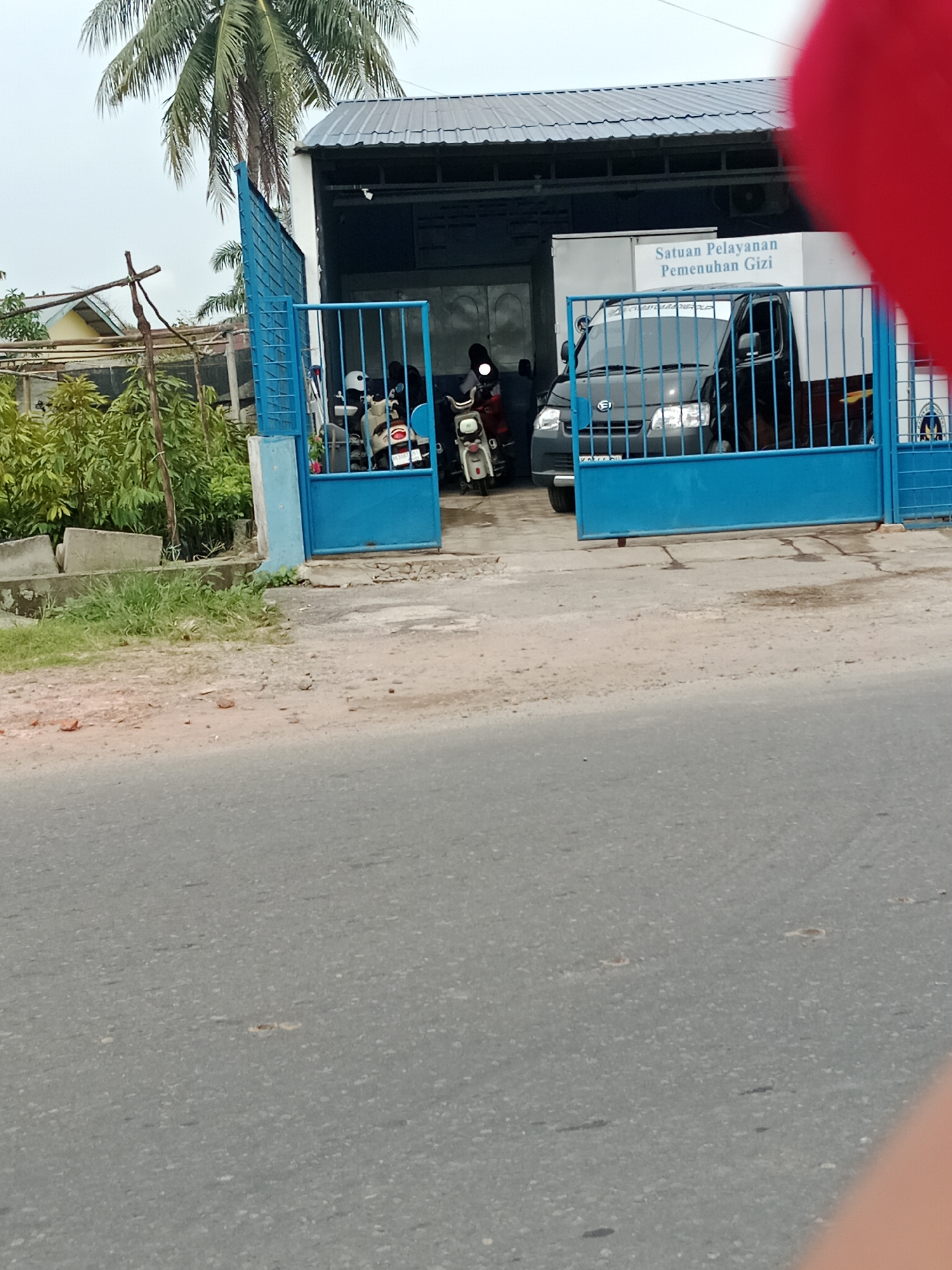 In Binjai, Indonesia, a grey pickup truck is parked behind a bright blue metal gate. A sign above the truck reads "Satuan Pelayanan Pemenuhan Gizi." Several motorcycles are parked inside the gate. A tall palm tree with green fronds is visible in the background. The scene is outdoors, with a paved road in the foreground and a building with a metal roof behind the gate.
