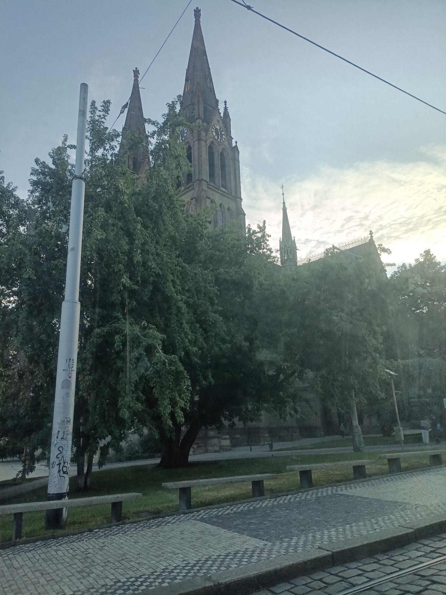 The image shows a view of a church with spires, partially obscured by trees. A lamppost stands in the foreground, on the left side of the image. The church appears to be made of stone and has a clock on one of its spires. The area in front of the church features a paved walkway with a checkered pattern and several benches. The sky is overcast, with some sunlight breaking through. The scene is likely an outdoor public space in a city. The specific location is the Church of Saints Peter and Paul in Vyšehrad, Prague, Czechia.