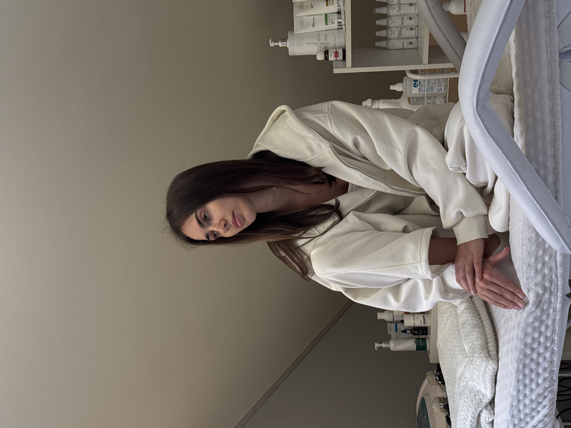 A young woman with long brown hair is sitting on a treatment bed in what appears to be a beauty or medical clinic. She is wearing a cream-colored hoodie. Her hands are resting on the bed which is covered with a white blanket. In the background, there is a shelf with various bottles and containers, likely skincare or treatment products. A piece of medical equipment can be seen in the lower part of the image. The scene appears to be indoors and well-lit. Location: Unknown, Unknown.