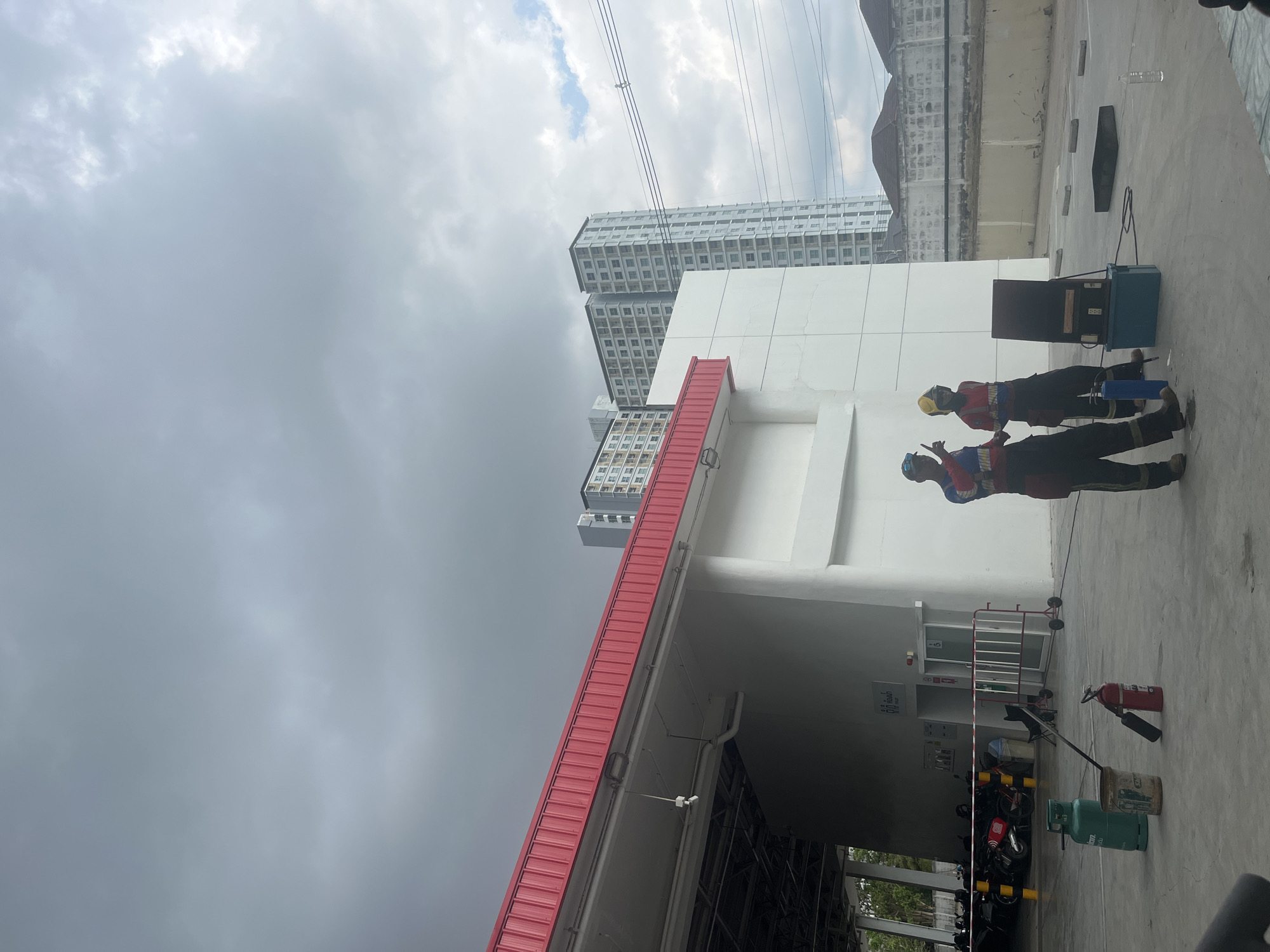 The image shows an outdoor scene in Pattaya City, Thailand. Two workers in red and yellow safety gear stand on a concrete surface near a white building with a red roof. Buildings are visible in the background. The sky is overcast. Tools, a fire extinguisher, and a green gas tank are nearby.