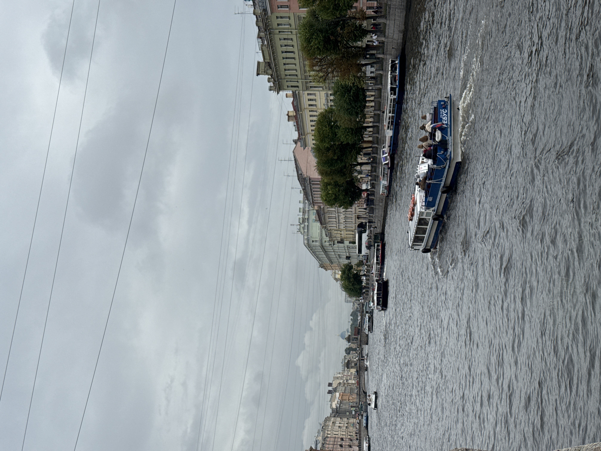 The image shows a canal in Saint Petersburg, Russia. The sky is overcast, indicating a cloudy day. Several boats are on the canal, including a passenger boat with people visible on its deck. Buildings line the canal, featuring a mix of architectural styles common in the city. Trees are planted along the canal banks, adding greenery to the urban landscape. The water in the canal is rippled, suggesting movement. Overhead, power lines are visible, crisscrossing the sky. On the passenger boat, the text "НЕБУС" is clearly visible. The scene appears to be a typical day of canal tours in the city, with the overcast weather contributing to a muted color palette.