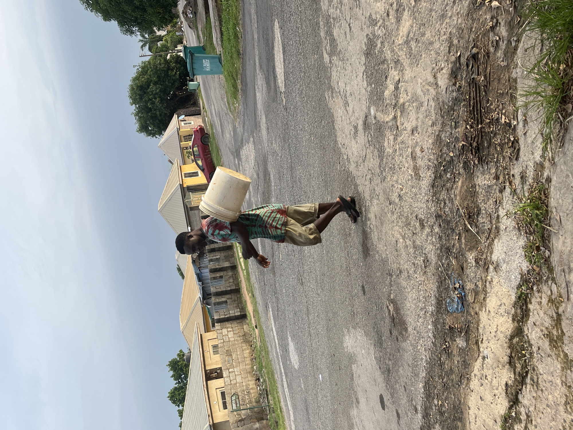 A man with dark skin walks on a paved road in Abuja, Nigeria, carrying a large, light-colored bucket in his right hand, slung over his shoulder. He wears a short-sleeved shirt with a colorful, abstract pattern of red, green, and blue stripes, and beige shorts. His feet are clad in dark sandals. The road is cracked and appears to be in disrepair, with debris and vegetation growing along its edges. In the background, several residential buildings with yellow and beige walls and silver-colored roofs are visible. A red car is parked on the side of the road. Two green trash bins, one with the words "HAPPY FAMILY" printed on it, stand near the road. The sky is overcast with a light gray hue. The overall scene depicts a common sight in an urban residential area.