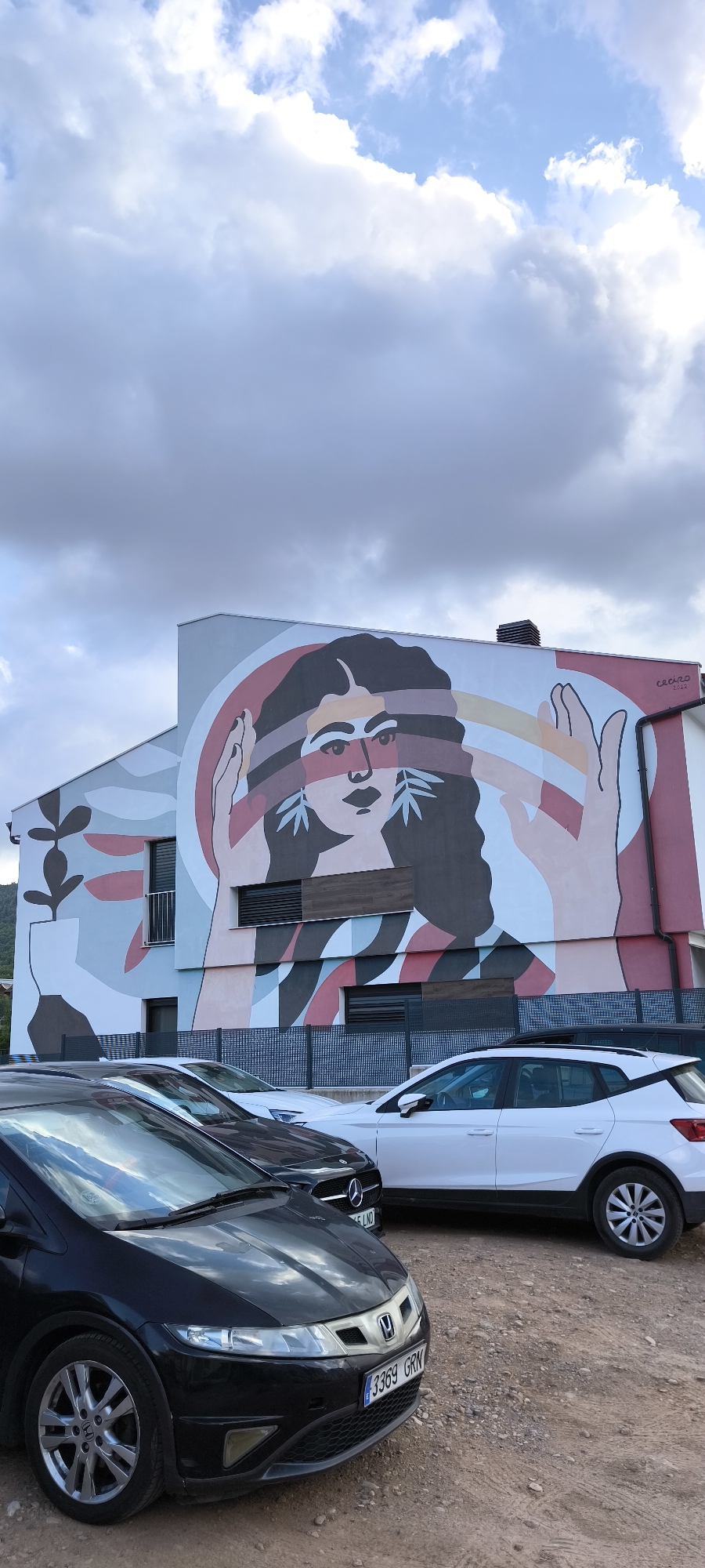 A vertical, eye-level, outdoor, daytime, wide shot captures a mural on the side of a building in Fanzara, Spain. The mural depicts a stylized woman with dark hair and a neutral expression. Her face is adorned with green leaf-like earrings and horizontal dark stripes across her eyes. Her hands are raised, with one hand partially obscuring her face. The mural uses a palette of pink, white, black, red, and light blue colors, with abstract shapes and lines forming a background. A white car is parked in front of the mural, to the right, and a black car is parked to the left. The ground is a gravelly, dirt surface. The sky above is overcast with large, white and gray clouds.