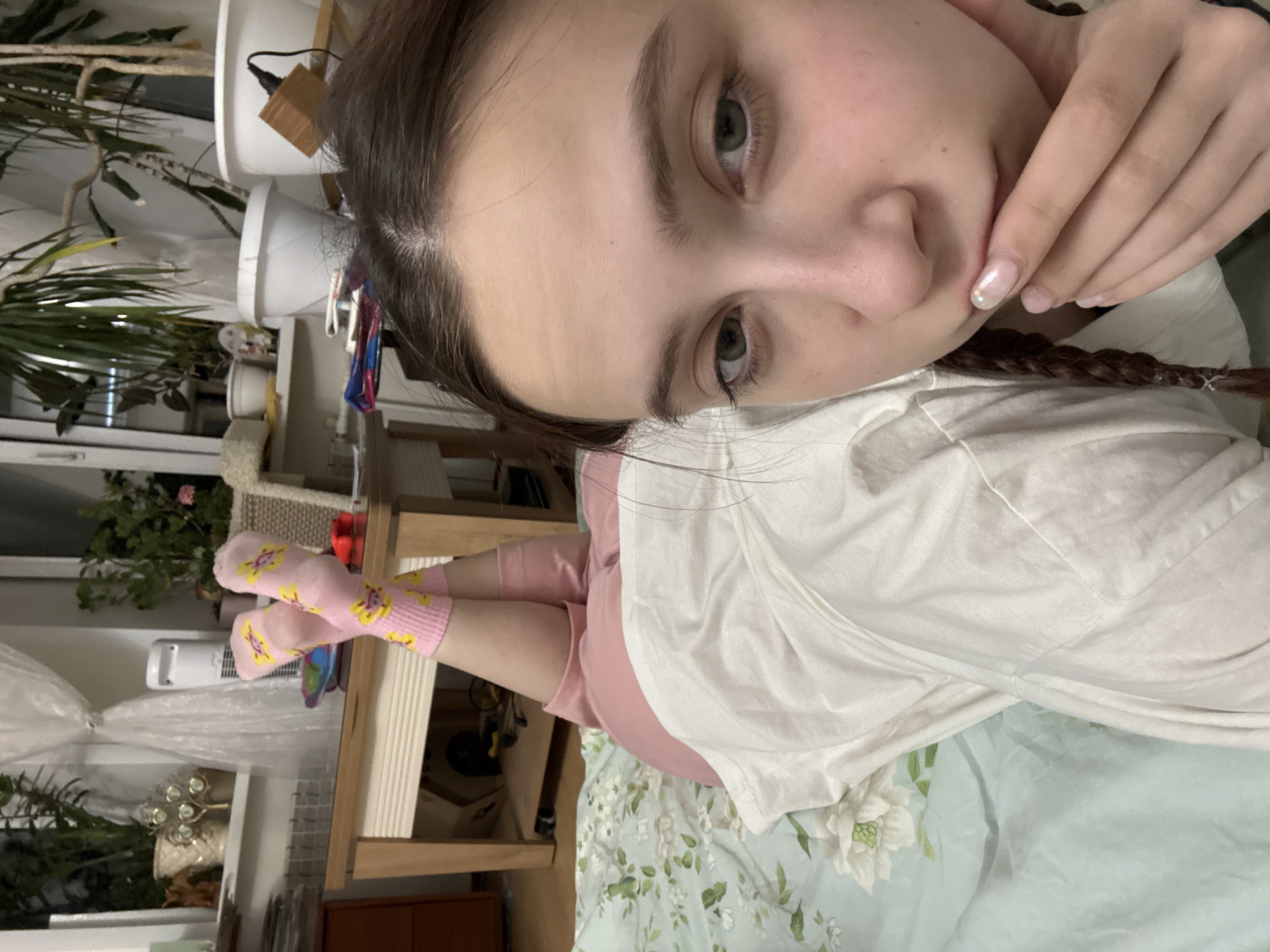 A selfie of a young woman indoors. She is lying on a bed with a light blue floral patterned bedspread. She is wearing a white t-shirt and light pink shorts. Her legs are stretched out, and she is wearing pink socks with yellow sun designs. Her hair is braided. A hand is touching her face. A wooden table or shelf is in the background. Beyond the table, there are plants, a window, and other household items. The scene does not contain location information that enables me to determine if it is within the city of Vogulka, Russia.