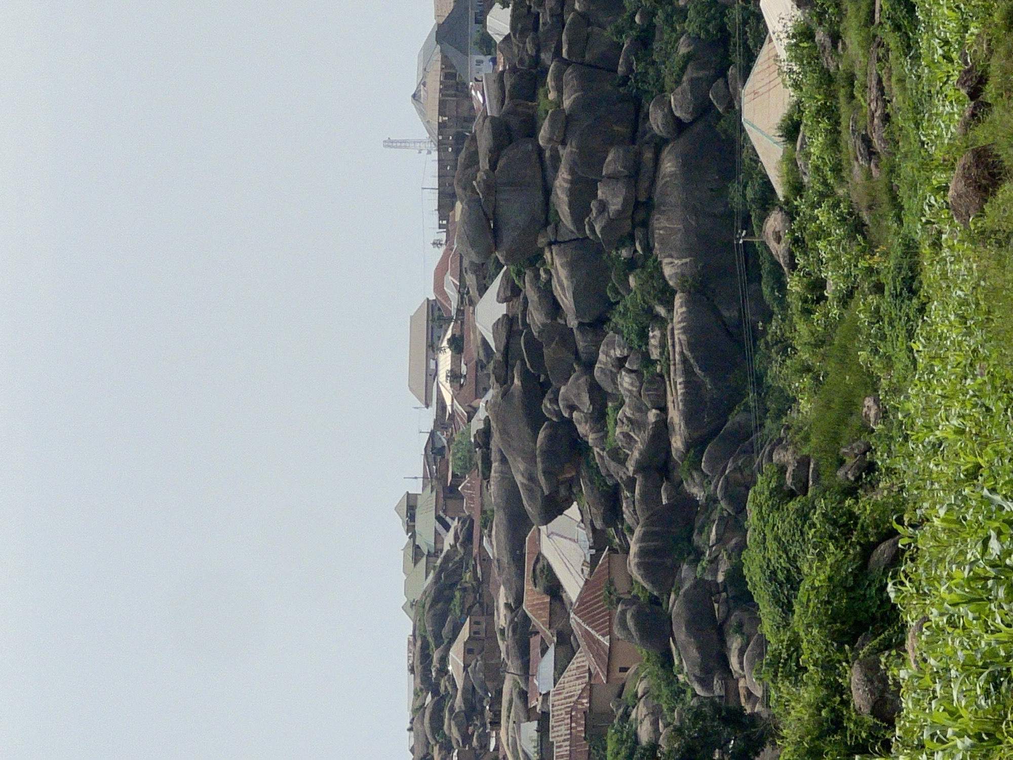 The image depicts a view of a settlement nestled among large rock formations, characteristic of the landscape in Jos, Nigeria. The foreground is filled with lush green vegetation, including what appears to be crops like corn, indicating an agricultural area. As the eye moves upwards, the terrain becomes increasingly rocky, with massive boulders dominating the scene. Scattered amongst these rocks are numerous houses with corrugated metal roofs and some with tiled roofs, suggesting a mix of traditional and more modern construction. The buildings are clustered together, following the natural contours of the hills. In the background, more buildings are visible, as well as a telecommunications tower, indicating a populated and developed area. The sky is overcast and pale, suggesting a hazy or cloudy day. There are no people or specific actions clearly depicted in the image, presenting a static scene of a rural or semi-urban settlement integrated with its natural rocky environment.