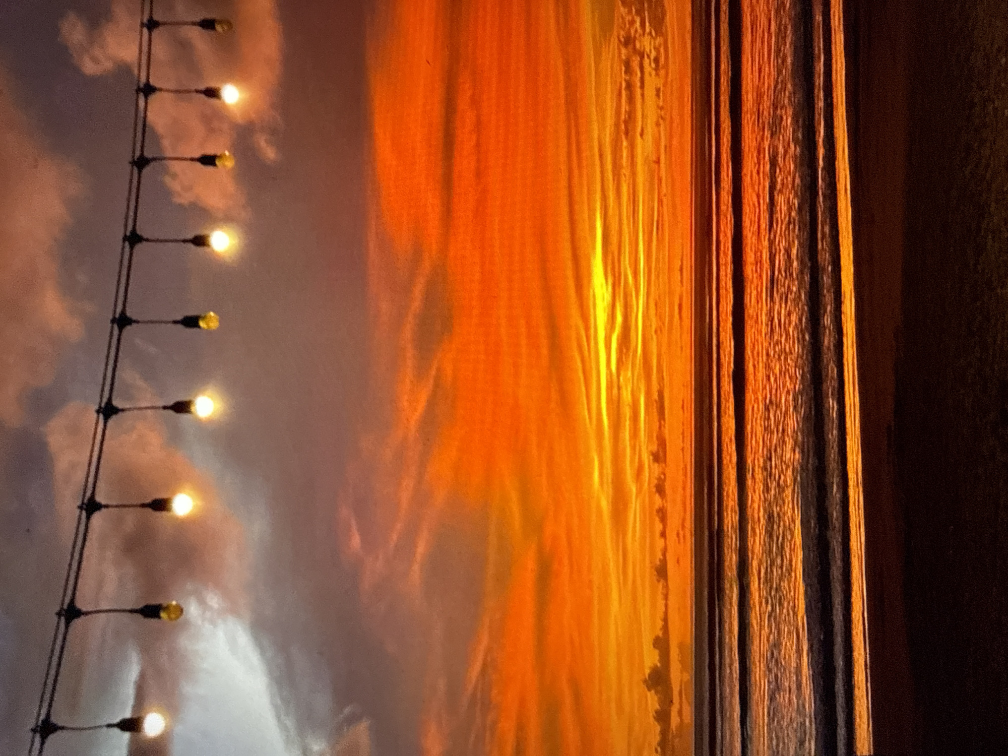 The image displays an expansive ocean sunset scene. A dark cable strung horizontally across the upper portion of the frame supports ten evenly spaced incandescent light bulbs, several of which are illuminated with a warm yellow to white glow, casting light downwards. The sky dominates the upper two-thirds of the image, transitioning dramatically from muted gray and soft purple clouds at the upper left to intense, fiery hues of red, orange, and bright yellow closer to the horizon, with layered clouds accentuating the vibrant colors of the sunset. Below the horizon, the ocean surface reflects the vivid sky, showing multiple distinct, parallel waves rolling towards the foreground. The closest waves exhibit some reflective whitecaps and streaks of orange light. The lowest portion of the image reveals a dark, silhouetted sandy beach. No individuals, specific man-made structures beyond the string lights, or discernible actions are present. Based on the visual evidence of an ocean and beach, this scene does not depict the inland city of Kardzhali, Bulgaria.
