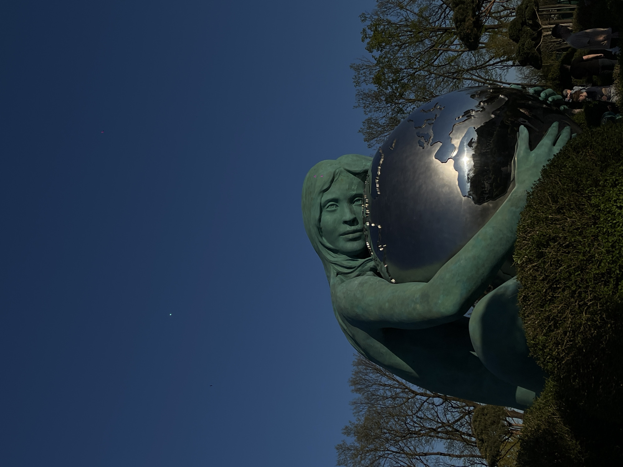 The image shows a sculpture of a female figure holding a globe against a clear blue sky. The figure is green in color, potentially made of bronze or a similar material, and appears to be squatting. The globe is made of a reflective material, like polished metal, and shows a distorted reflection of the surrounding environment. Parts of trees and bushes are visible around the sculpture. In the background, indistinct figures can be seen, implying a public space. While the location is identified as Krasnodar, Russia, the context lacks detail to confirm the exact location.