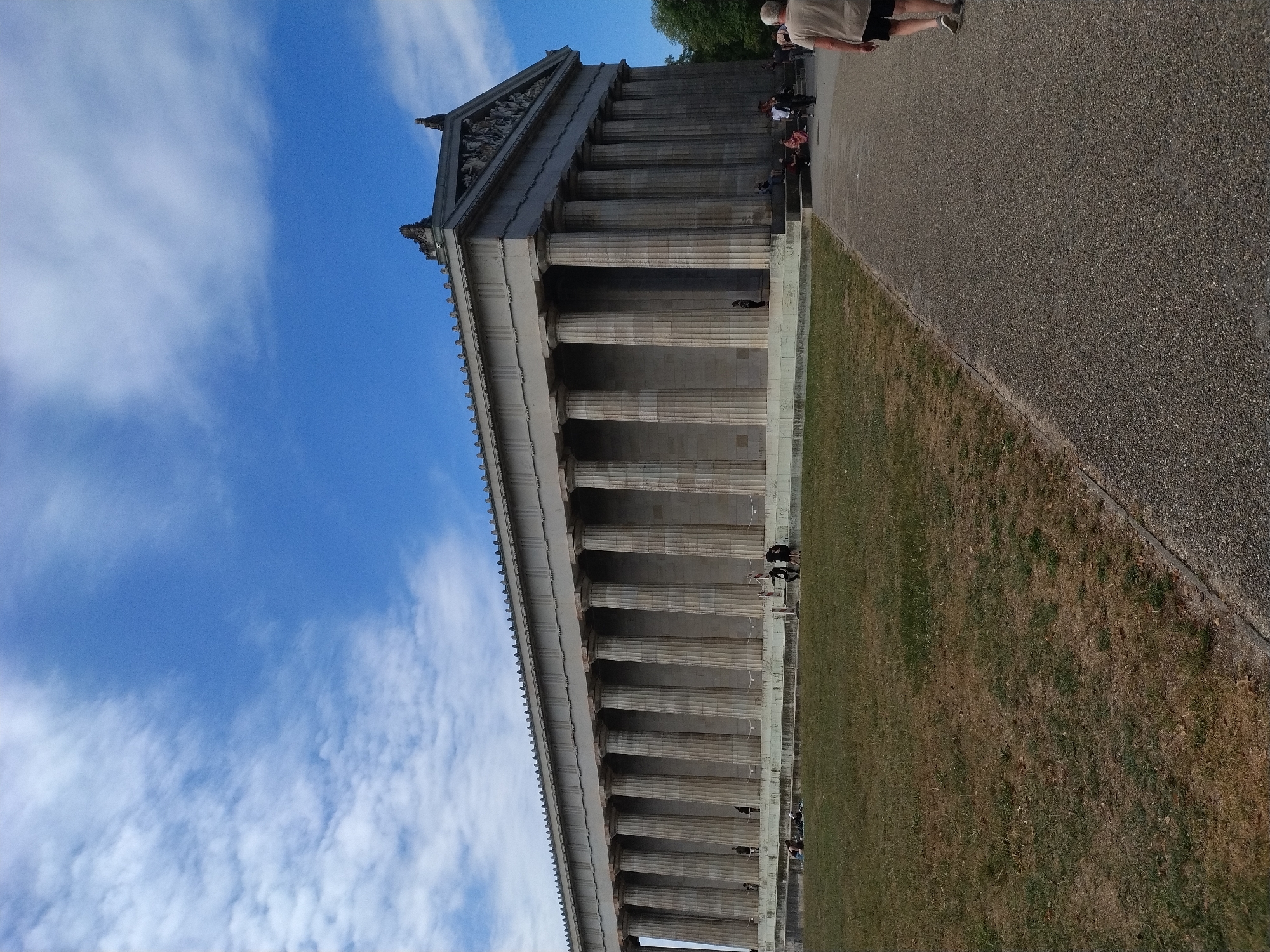 The image depicts the Walhalla memorial, a majestic neoclassical building located on a hill overlooking the Danube River in Donaustauf, Germany. The building is characterized by its long colonnade of Doric columns, a pediment adorned with sculptures, and a grand structure suggesting historical significance. In the foreground, a paved walkway curves around a patch of dry, yellowish-brown grass. Several individuals are visible; some are walking along the path, while others are seated or standing on the grassy area or the steps leading up to the building. The sky is a bright blue with scattered white clouds, suggesting a clear day. The overall scene conveys a sense of grandeur and historical preservation, with visitors enjoying the site.