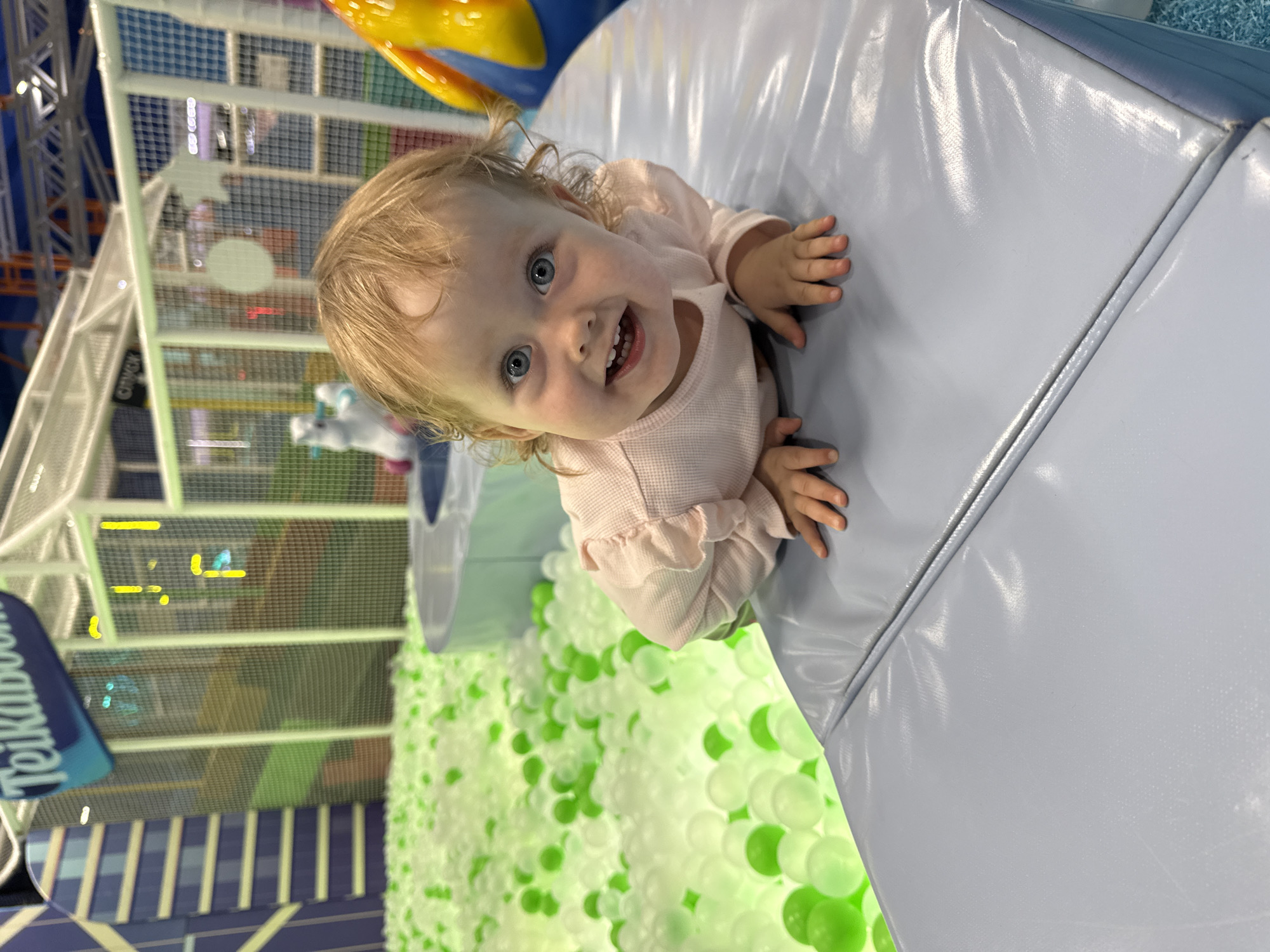 A young child with blonde hair and blue eyes is positioned on a light blue padded surface in what appears to be an indoor playground. The child is wearing a light pink long-sleeved shirt and is smiling with visible teeth. Behind the child is a ball pit filled with green and white balls. A structure is visible in the background, with a sign that reads "Teikaboom." The specific location is not identified in the image.