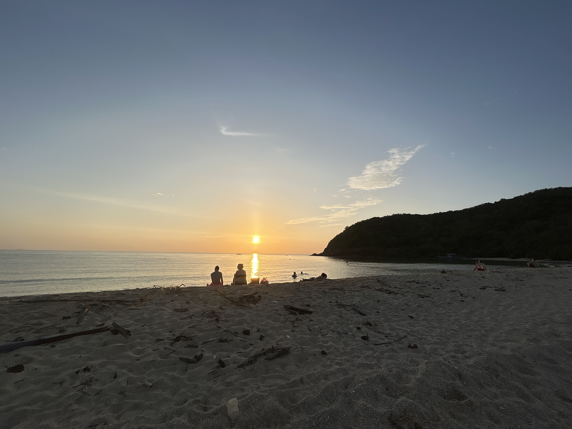 This is a horizontal, eye-level, medium shot of a beach at sunset in Ko Pha-ngan District, Thailand. The sun is setting over the calm ocean, casting a warm glow across the sky and water. The sand in the foreground is a dark brown, with scattered driftwood. In the middle ground, there are three people sitting on the sand facing the ocean, with a fourth person lying down to their right. In the background, a dark green, tree-covered hill rises from the water. A small boat is visible in the distance. The sky is a gradient of pale blue to orange, with a few wispy clouds.