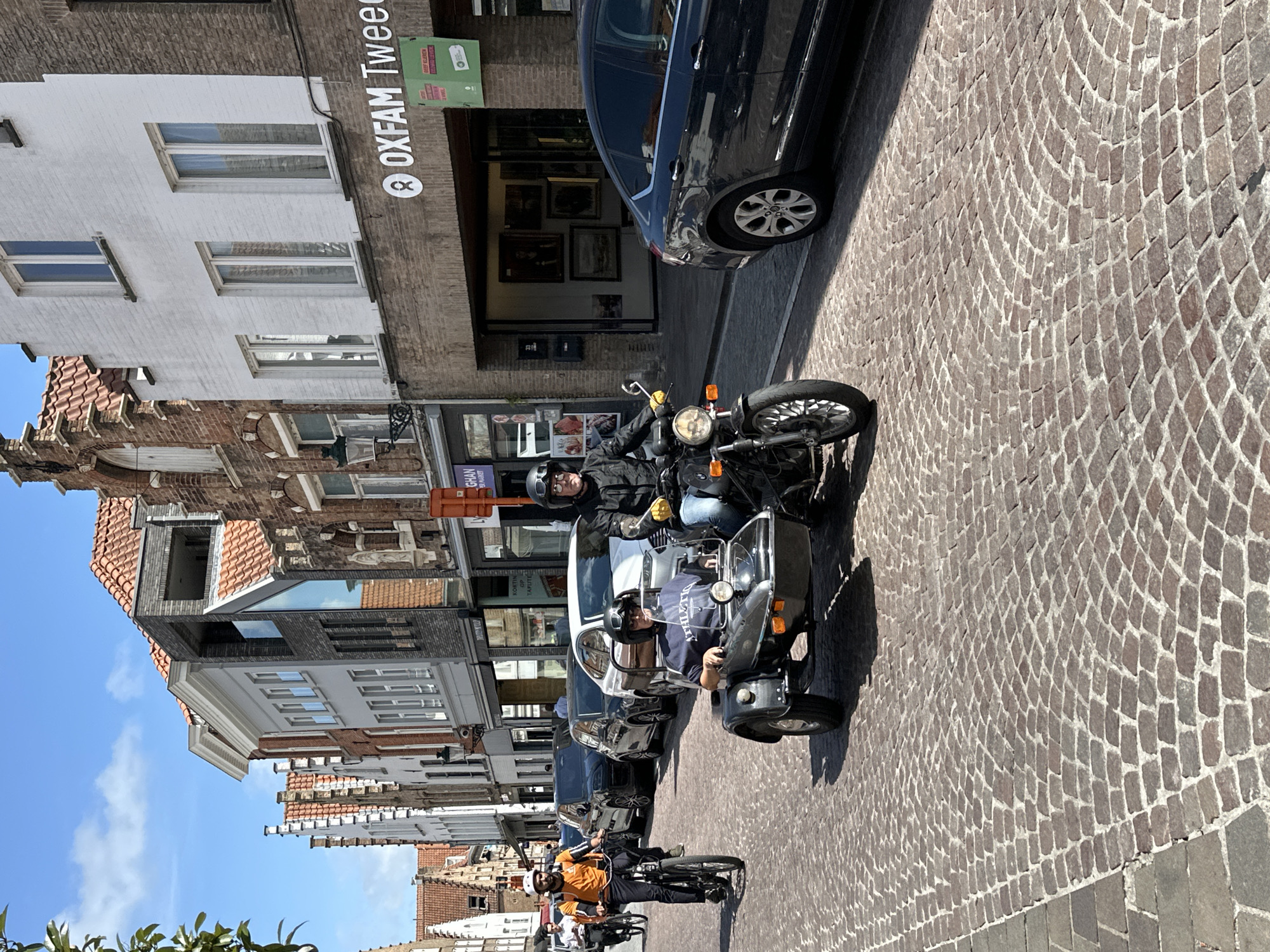 The image depicts a street scene in Bruges, Belgium. In the foreground, a man wearing a helmet and dark clothing rides a motorcycle with a sidecar. A passenger, also wearing a helmet and a dark t-shirt with "ATHLETIC" printed on it, sits in the sidecar. The motorcycle and sidecar are traveling on a cobblestone street lined with buildings on both sides.
To the left of the motorcycle, another person wearing an orange shirt and helmet is riding a bicycle. In the background, several cars are parked along the street. The buildings are characteristic of Bruges' architecture, with red-tiled roofs and multi-story facades. One building on the right has a sign that reads "OXFAM Tweedehands". The sky is blue with some clouds, suggesting a clear day. The scene appears to be a typical day in a European city with both pedestrian and vehicular traffic.