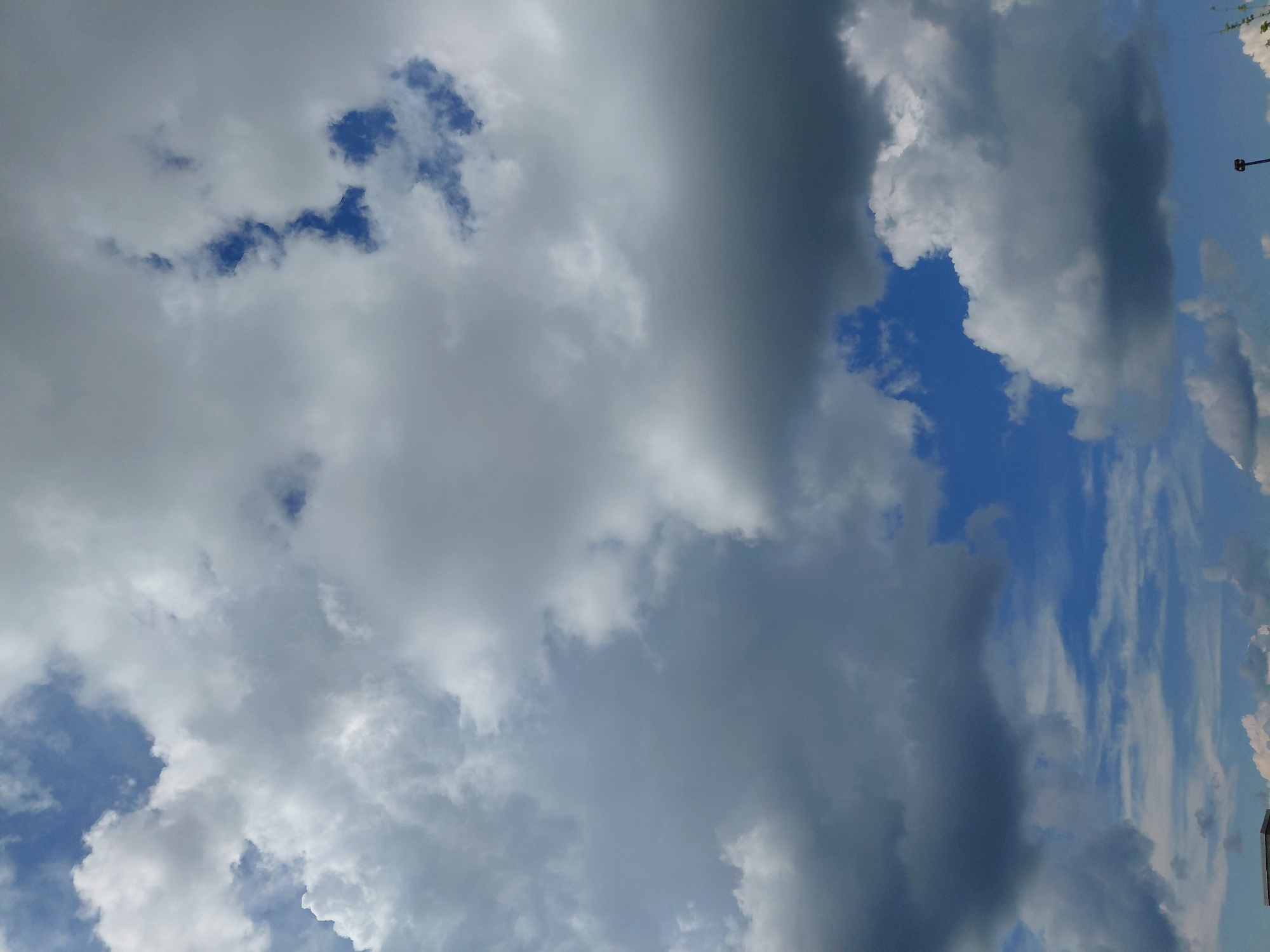 The image shows a cloudy sky with patches of blue visible. The clouds are varied in texture and density, with some appearing fluffy and white, while others are darker and more ominous. The overall impression is that of a dynamic sky, possibly indicating changing weather. In the lower portion of the image, a sliver of a building is visible, suggesting an urban or suburban environment. The presence of a lamppost in the lower right corner further reinforces this interpretation. The lighting suggests it is daytime, with the sun possibly obscured by the clouds, leading to diffuse illumination. There are no people or specific actions depicted in the scene. The sky takes up the majority of the frame, with the ground-level elements forming a small part of the composition. The location is specified as the city of Lexington, United States of America.