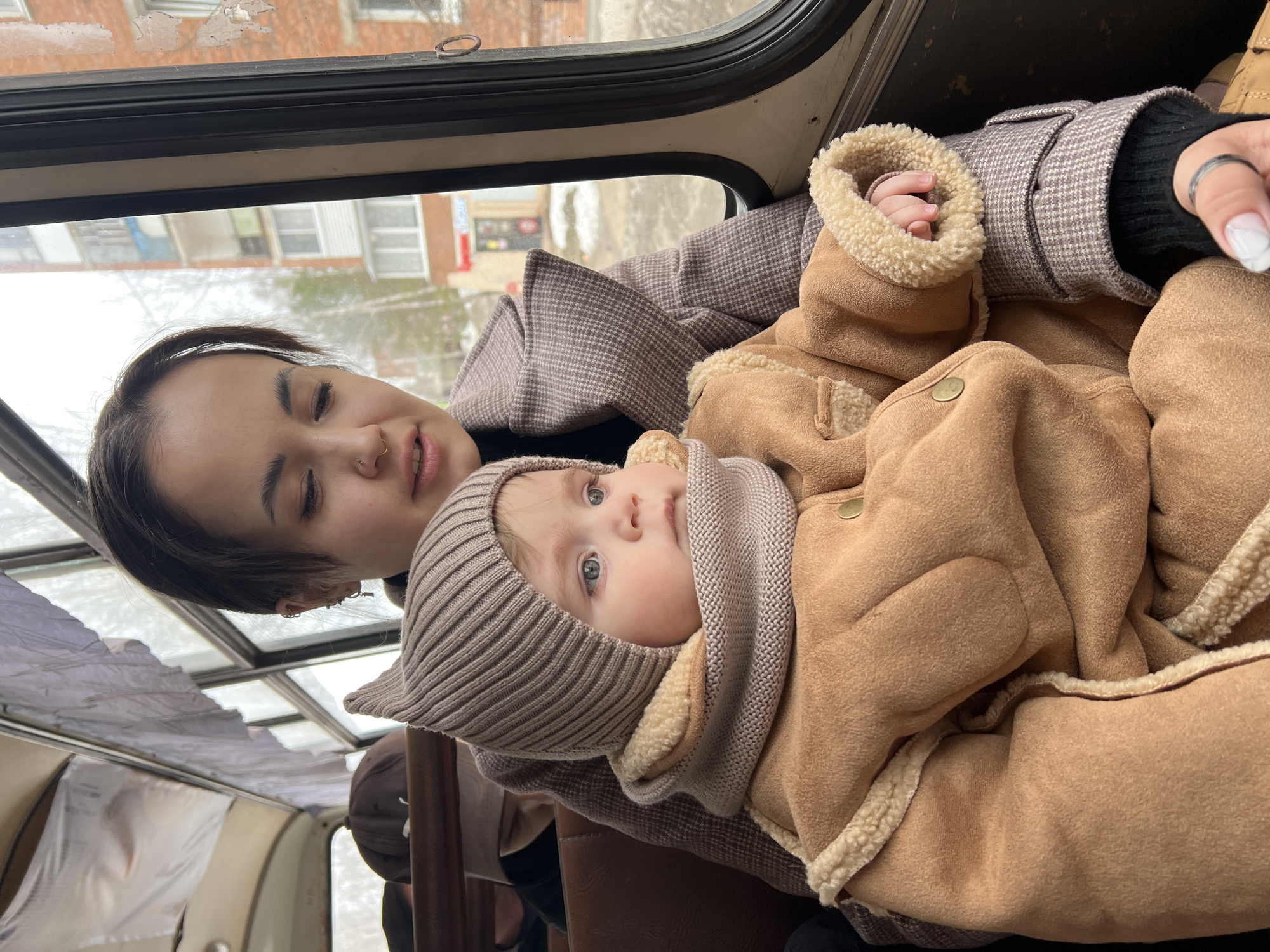 A woman is holding a baby inside a vehicle, likely a bus, with a view of the city of Noviy, Russia, visible through the window.
The woman has dark hair pulled back, a gold nose ring, and is wearing a brown and white checkered coat over a black sweater. She is looking at the baby in her arms with a soft expression.
The baby is bundled in a brown, sheepskin-lined snowsuit and a knitted hood, and scarf. The baby is looking up with wide, bright eyes. The baby's hand is partially visible in the woman's hand.
The interior of the bus has brown seats. Through the bus window, it's possible to see the reflection of the city. In the background, there is a brick building visible. The scene suggests it is a cold day.