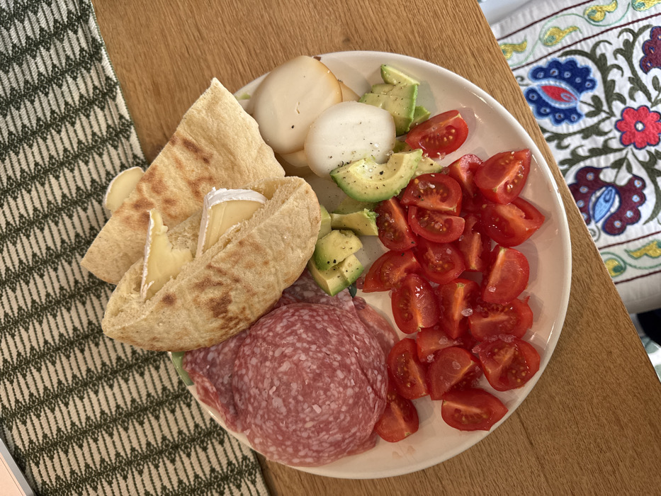 The scene depicts a meticulously arranged platter of food on a light-colored, oval plate, positioned on a wooden table surface within an indoor setting in Lecce, Italy. The plate contains several distinct food groups. On the left, a mound of thinly sliced, light red cured meat, possibly salami, with visible fat speckles, is present. Adjacent to this, towards the center-top, are several pieces of light brown flatbread, some folded to reveal slices of creamy yellow cheese with white rinds. Below the flatbread and cheese, a scattering of green avocado chunks, seasoned with black pepper, and white, irregularly shaped cheese pieces, one appearing slightly darker or smoked on an edge, occupy the right side of the plate. The bottom left section of the plate is filled with numerous bright red cherry tomatoes, quartered or halved, displaying their inner seeds and sprinkled with coarse salt crystals. The wooden table exhibits two distinct fabric runners: one with a dark green geometric pattern in the upper left background and another with a colorful floral and paisley embroidery pattern in red, blue, and green visible in the lower right foreground. No individuals are present in the image.