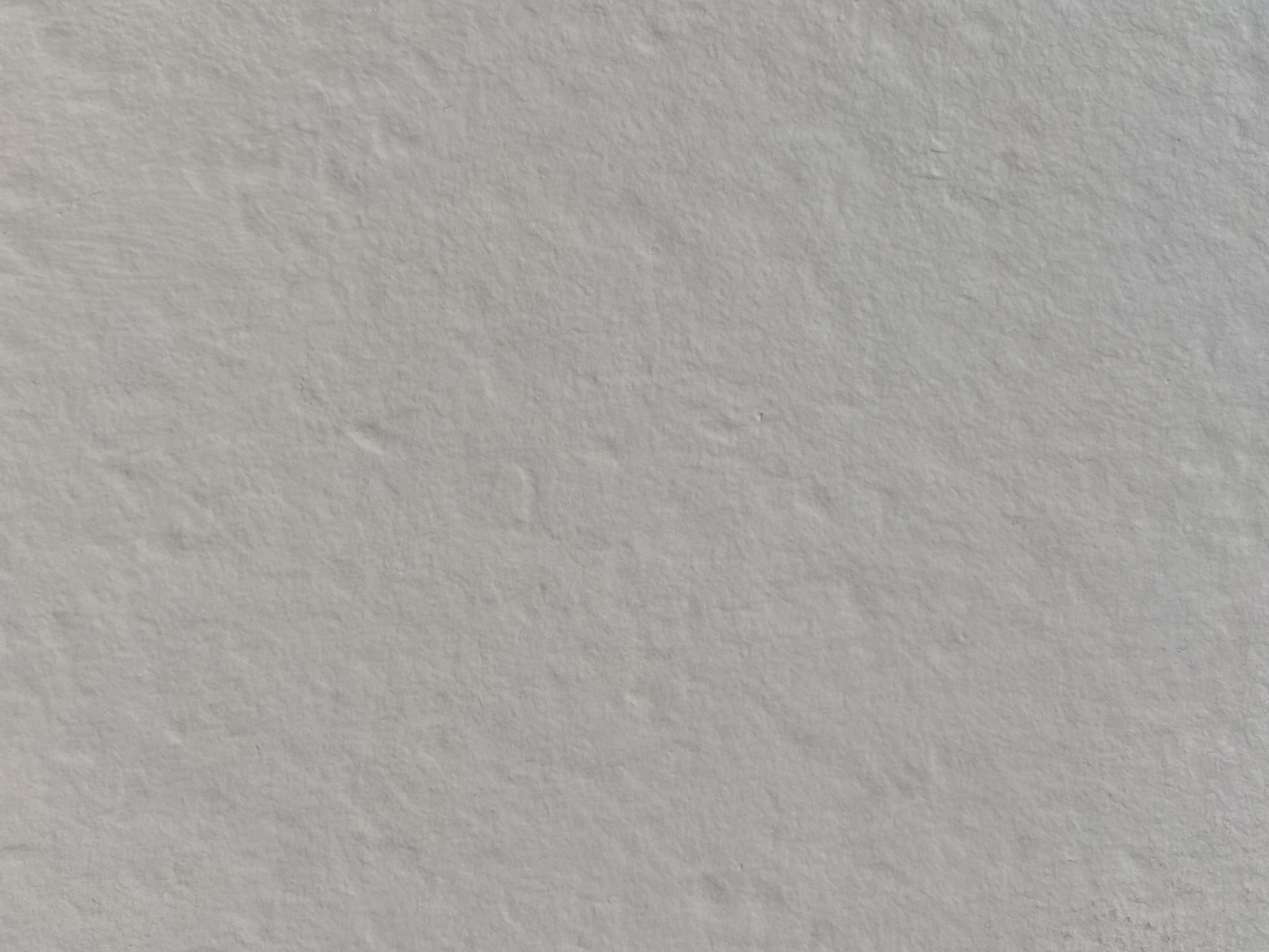 The image shows a close-up view of a textured, light gray surface. The texture appears to be rough and uneven, with small bumps and indentations visible throughout. There are no discernable objects, people, or actions captured in the frame. The lighting is even, casting no distinct shadows. The overall impression is of a neutral, abstract background. The location is unknown.
