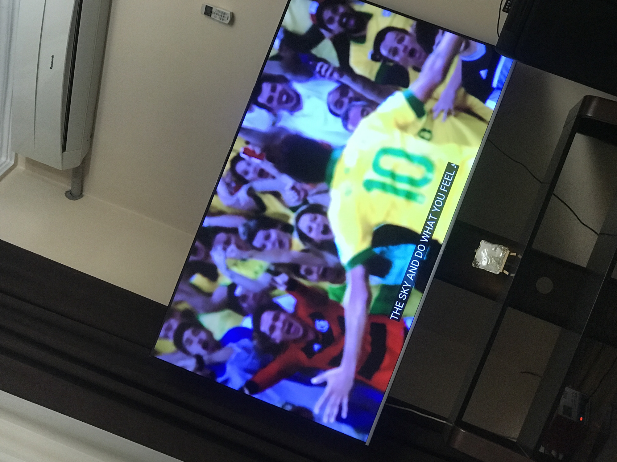 The image shows a television screen displaying a soccer game with fans and a player wearing a yellow jersey with the number 10 on it. The television is placed on a black stand. A white air conditioner is mounted on the wall above the television. There is also a remote control visible on the wall. The location is unspecified.