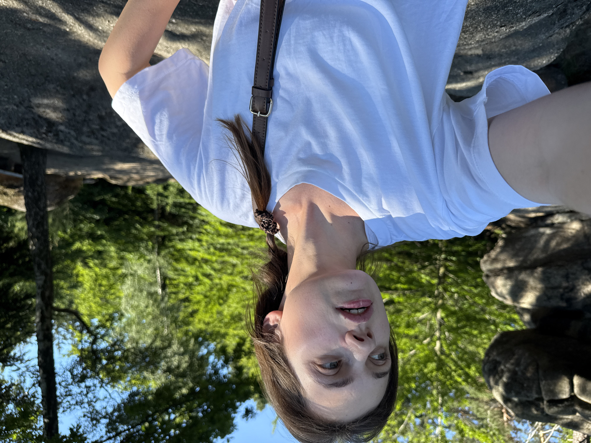 A person is captured in a close-up, upside-down selfie, with the background suggesting an outdoor, natural setting. The subject, a person with long brown hair tied in a braid, is wearing a white t-shirt and a brown strap across their shoulder. Their face is turned towards the camera, with eyes open and a slightly parted mouth, conveying a relaxed or engaged expression.
The background is dominated by lush green foliage, characteristic of a forest or park, with sunlight filtering through the leaves, indicating it's daytime. Large grey rocks are also visible, hinting at a landscape that might include rocky terrain or outcrops. The overall impression is one of being outdoors in a bright, possibly sunny environment, with nature as the primary backdrop. There is no visible text in the image. The location context of Szklarska Poręba, Poland, suggests a mountainous or forested region.