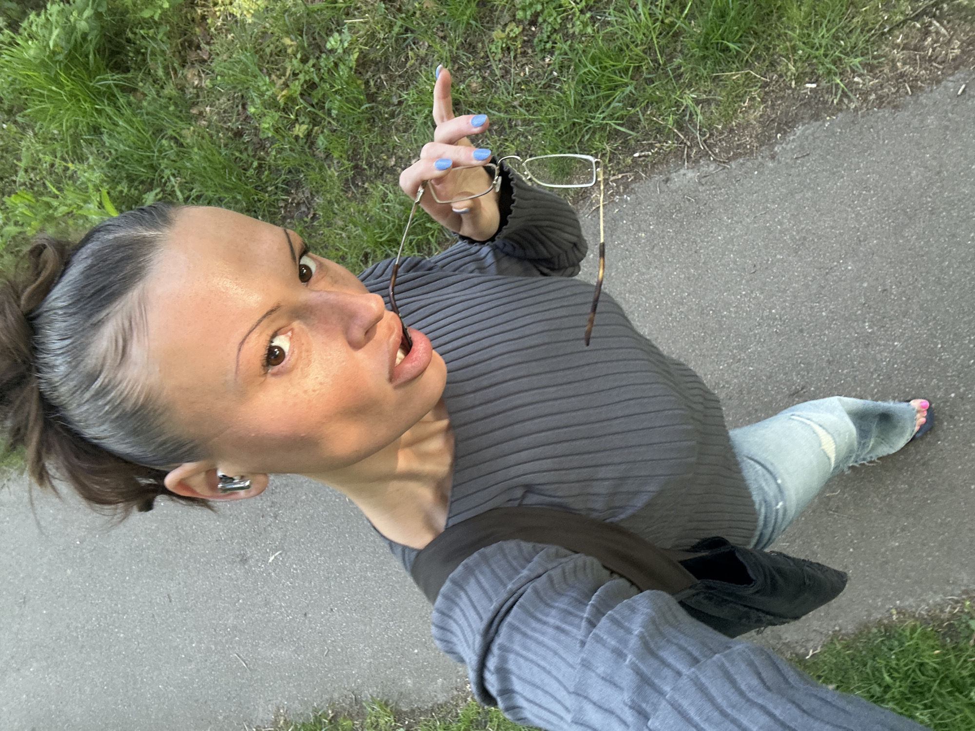 A woman is taking a selfie outdoors, likely in Tunbridge Wells, UK. The woman has brown hair pulled back in a bun and is wearing a gray, ribbed long-sleeved top and ripped blue jeans. She is standing on a paved path with grass on either side. She holds eyeglasses in her right hand, with light blue nail polish on her fingernails. A black bag hangs on her left shoulder. She wears an earring in her left ear. One foot is visible with a pink painted toenail.