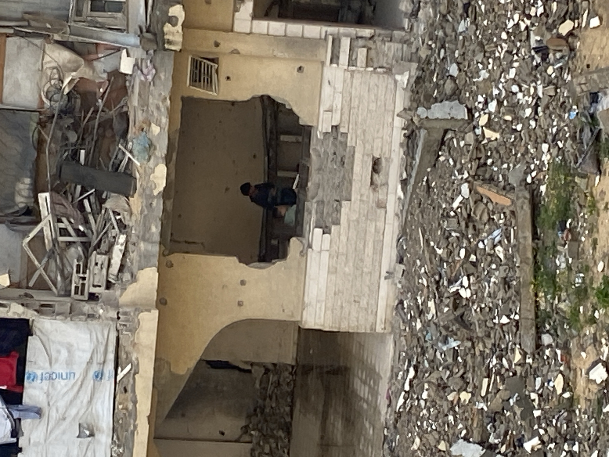 The image depicts a severely damaged multi-story building in Khan Yunis, Palestinian Territory, surrounded by extensive rubble. On the top visible floor, a white sheet or tarp bearing the "unicef" logo hangs vertically from an exposed section, with white and red clothing items draped nearby. The floor below this exhibits significant structural collapse, exposing a dense accumulation of twisted rebar, shattered concrete, and general debris.
At a lower level, a large section of an exterior wall is missing, creating an open-air view into a room. Inside this room, two individuals are present. One individual, wearing dark clothing, stands upright, appearing to observe or interact with something on a low, horizontal structure. Behind and slightly to the left of the standing person, a second individual, wearing a light-colored top, is seated or crouched on a similar low structure. The remaining interior walls of this exposed room are beige and show multiple small circular impact marks, consistent with bullet holes. A louvered vent is visible on the upper right wall section. The lower part of this building section features a partially intact wall constructed from light-colored, horizontal blocks. To the left of the exposed room, a dark arched opening reveals a reflective, possibly water-covered, floor.
The foreground and immediate surroundings of the building are covered by a vast, uneven expanse of gray and beige rubble, composed of fragmented concrete, stone, and other debris, with sparse patches of green vegetation emerging intermittently.