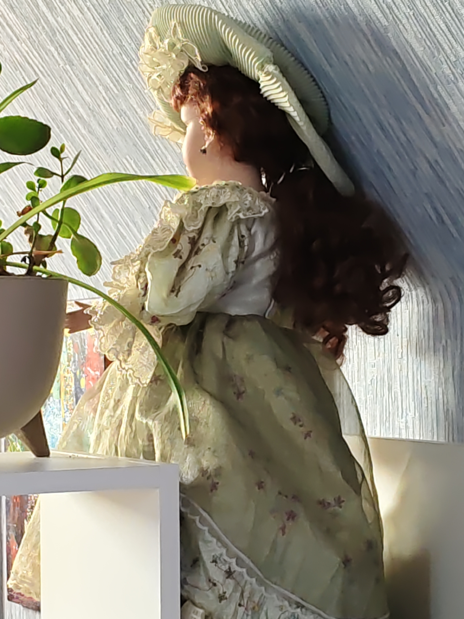 A doll with auburn curly hair and a pale complexion is positioned in profile, facing left. It wears an elaborate pale green dress with ruffled lace trim at the neckline and sleeves. The dress is adorned with a floral pattern of small purple flowers. A wide-brimmed, pale green hat with pleated details and a decorative ribbon sits atop its head. The doll is placed in front of a pale blue, textured wall, with a plant in a light gray pot to its left. The lower portion of the image shows a white shelf or furniture item. The scene appears to be indoors, possibly in a residential setting. There are no visible indications of the doll's location within the city of Unknown, Russia.