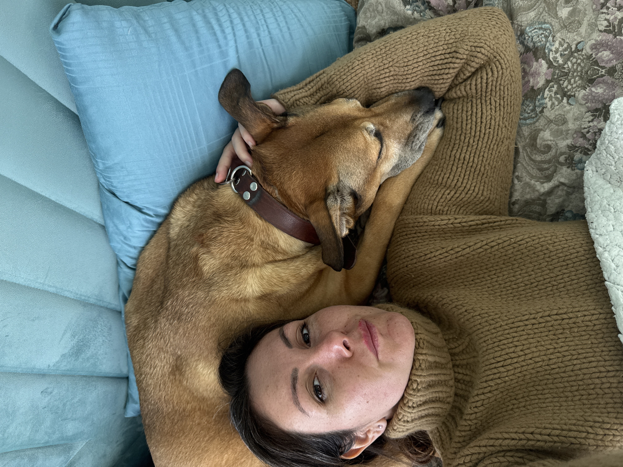 A woman and a dog are lying together on a couch. The woman is on her back, facing the camera. She has fair skin, dark hair, and a neutral expression. She is wearing a brown turtleneck sweater. The dog, which appears to be a mixed breed with brown fur, is lying on its side with its head resting on the woman's arm. The dog is wearing a brown leather collar. They are both on a blue cushion. The couch has a floral print. The location is unknown, and there is no indication of Byala Slatina, Bulgaria.