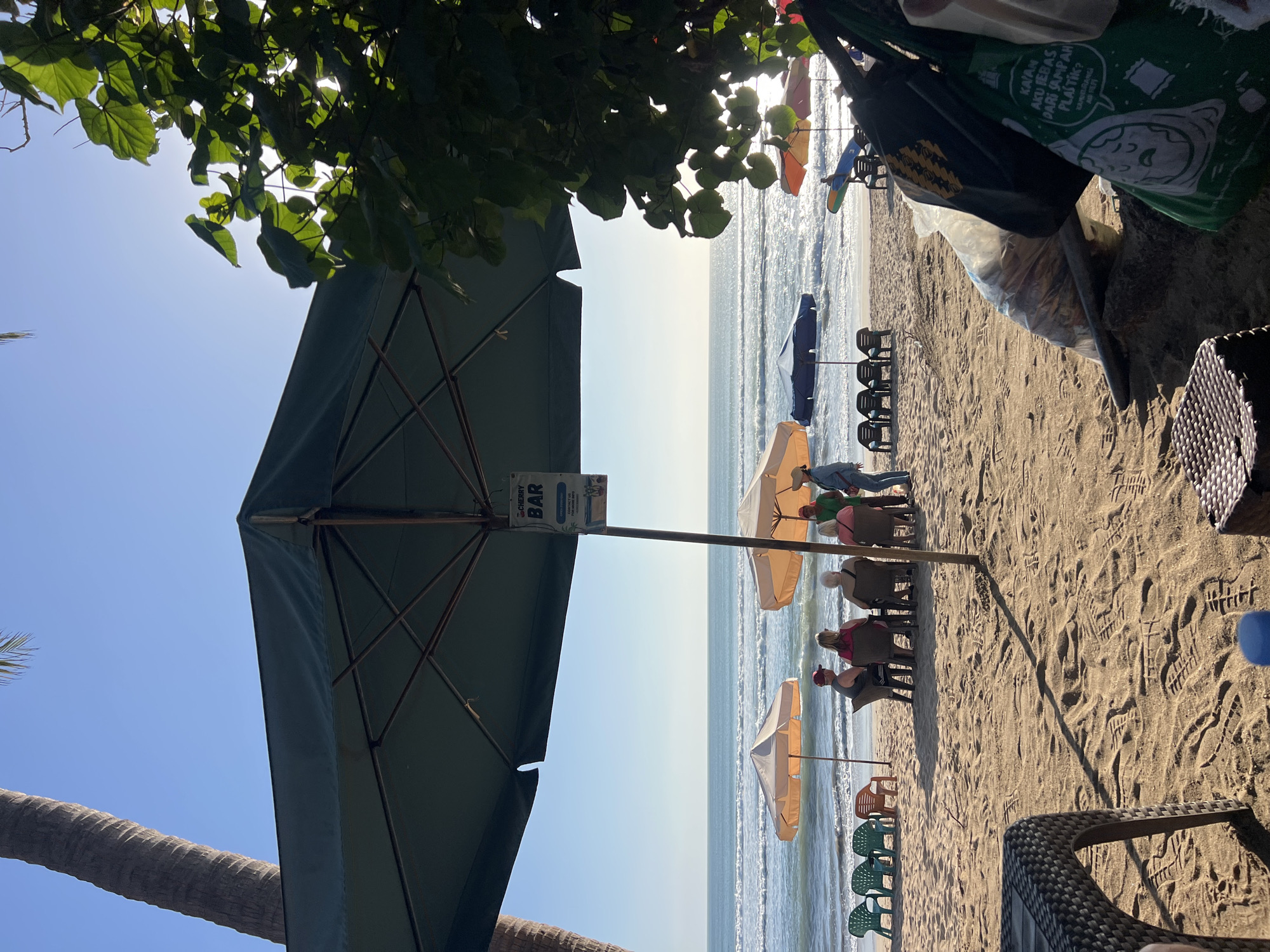 This image depicts a beach scene in Kuta, Indonesia, under a clear blue sky. A large, dark green umbrella is prominently positioned in the foreground, casting a shadow on the sandy ground. Beneath this umbrella, a group of people are seated at tables, facing the ocean. They appear to be engaged in conversation or relaxing.
Further back, closer to the water, several smaller, lighter-colored beach umbrellas dot the sand, with some beach chairs placed beneath them. The ocean is calm with gentle waves rolling towards the shore. In the distance, the horizon is visible, separating the sea and the sky.
To the right of the frame, lush green foliage from a tree hangs over the scene, its leaves dappled by sunlight. Some bags are visible in the foreground, including a green bag with text and imagery. The sand is textured with footprints, indicating recent activity. A palm tree trunk is visible to the left, adding to the tropical ambiance. The overall impression is a relaxed and sunny day at a beach resort.