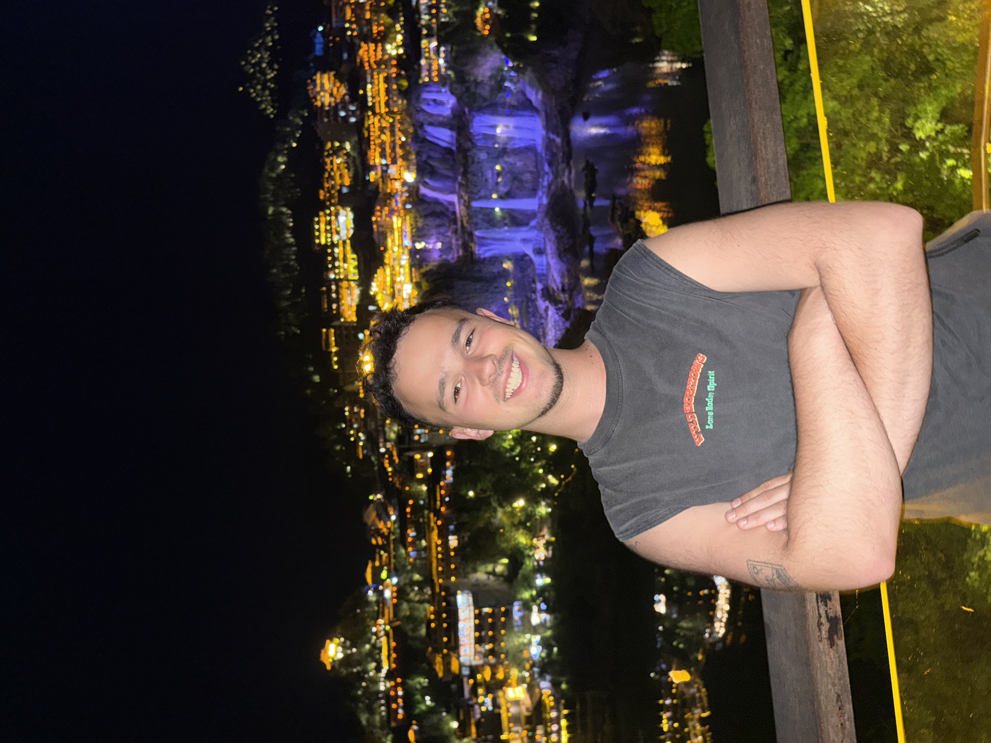 A man smiles at the camera, arms crossed, standing on a balcony. He wears a gray t-shirt. Behind him is a night view of the illuminated Furong Ancient Town in Hunan Province, China, including waterfalls and buildings lit up with yellow and blue lights. The scene is captured at night, with a dark sky above the town. A wooden railing is visible on the right side of the frame.