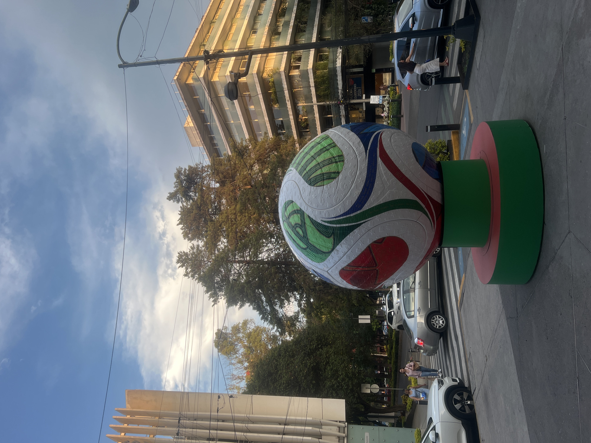The image shows a large, stylized soccer ball sculpture in Mexico City, Mexico. The ball rests on a cylindrical base painted green and red, the colors of the Mexican flag. The ball itself is white with decorative patterns in green, blue, and red. The background features a multi-story building with glass windows and greenery on the balconies. Trees and a clear, blue sky with scattered clouds are visible behind the sculpture and the building. Cars are parked along the street, and a few people are walking nearby. Streetlights and power lines are also present in the frame.