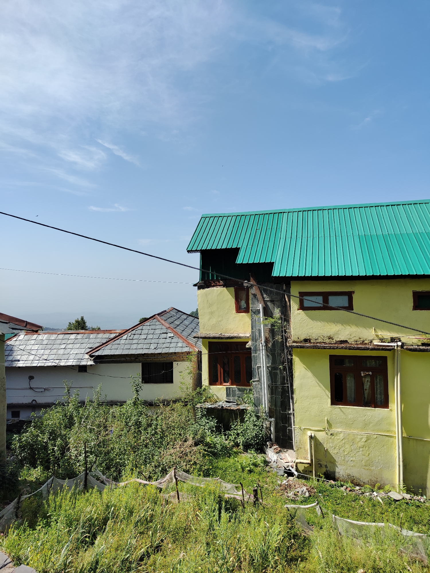 The scene depicts a residential area in Haini, India. A two-story yellow building with a green corrugated metal roof dominates the right side of the frame. The building has dark brown window frames. Adjacent to it on the left is another building with a gray slate roof. Overgrown vegetation covers the ground in front of the buildings. A wire stretches across the upper part of the image. The sky is visible above the buildings and shows a partly cloudy day.