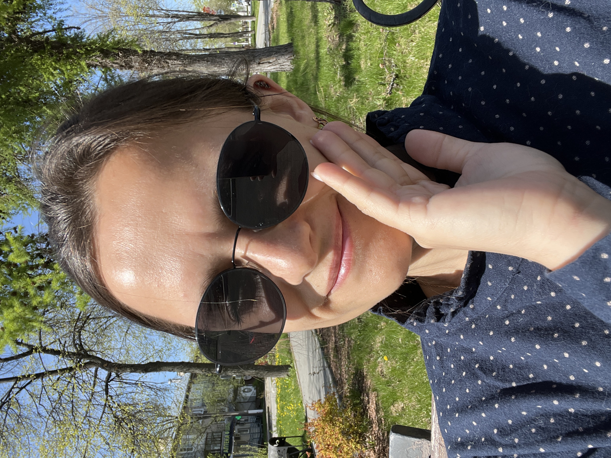 A woman wearing sunglasses and a polka-dotted blue dress is sitting outside on a sunny day. The location is Chaikovsky, Russia based on the sign "АПТЕКА" partially visible in the background. The woman is touching her face with her right hand. Trees are visible in the background along with a building. A green lawn area and sidewalk are visible between the trees and the building.