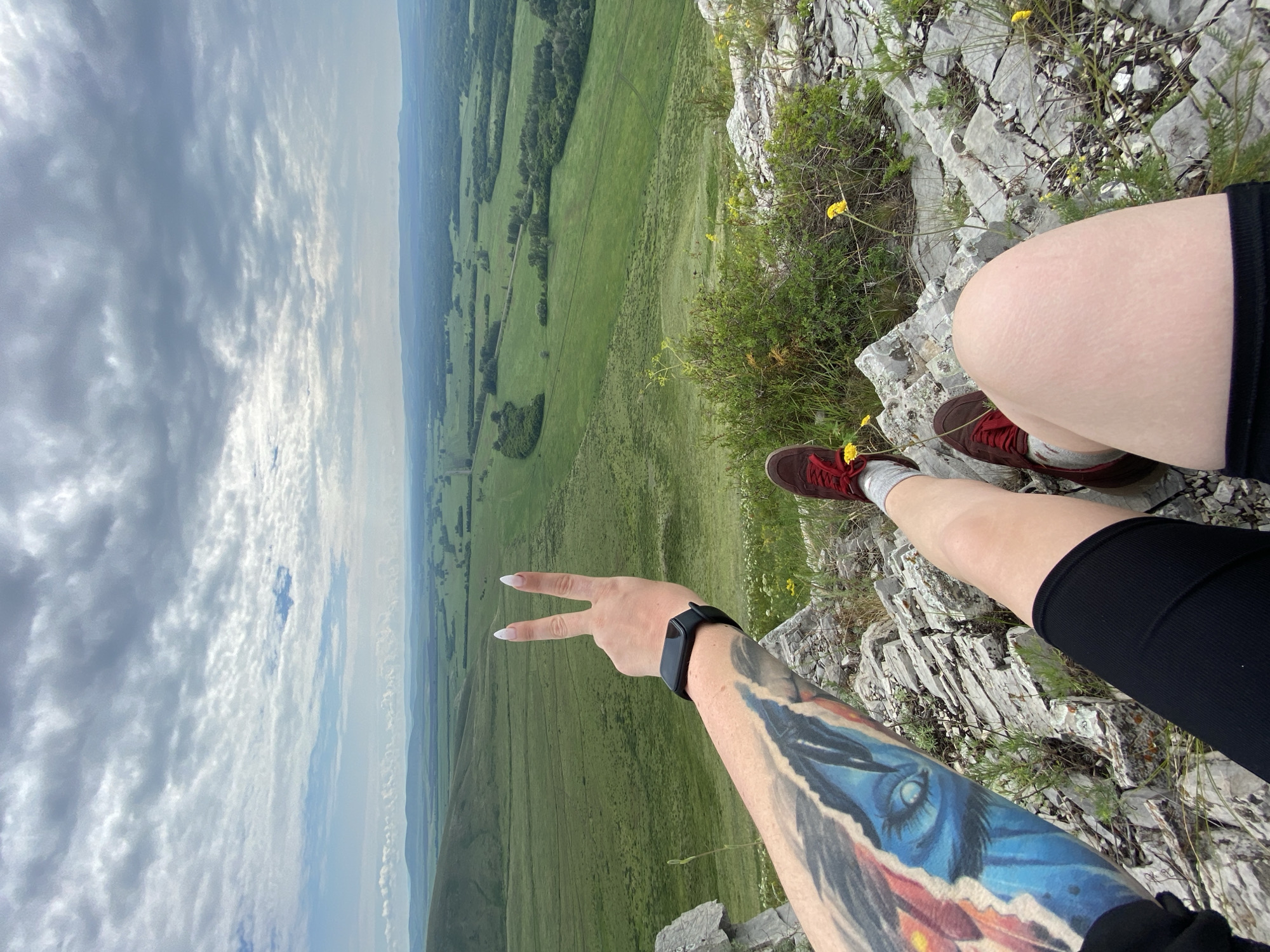 A person's arm and legs are visible in the foreground, positioned on rocky terrain overlooking a vast, green valley under a cloudy sky. The person's left arm, adorned with a colorful tattoo of an eye and abstract shapes, is raised, displaying a peace sign with their fingers. A black fitness tracker is worn on their wrist. Their right leg is bent, with a red sneaker visible. The terrain consists of grey rocks with sparse green vegetation and small yellow wildflowers. The valley stretches into the distance, featuring fields, trees, and rolling hills. A road or path is faintly visible winding through the landscape. The sky is filled with a mix of light and dark grey clouds, with hints of blue peeking through. The image appears to be taken from a high vantage point, suggesting the person is on a hilltop or mountain. The location is specified as Verkhnyaya Chernorechka, Russia.