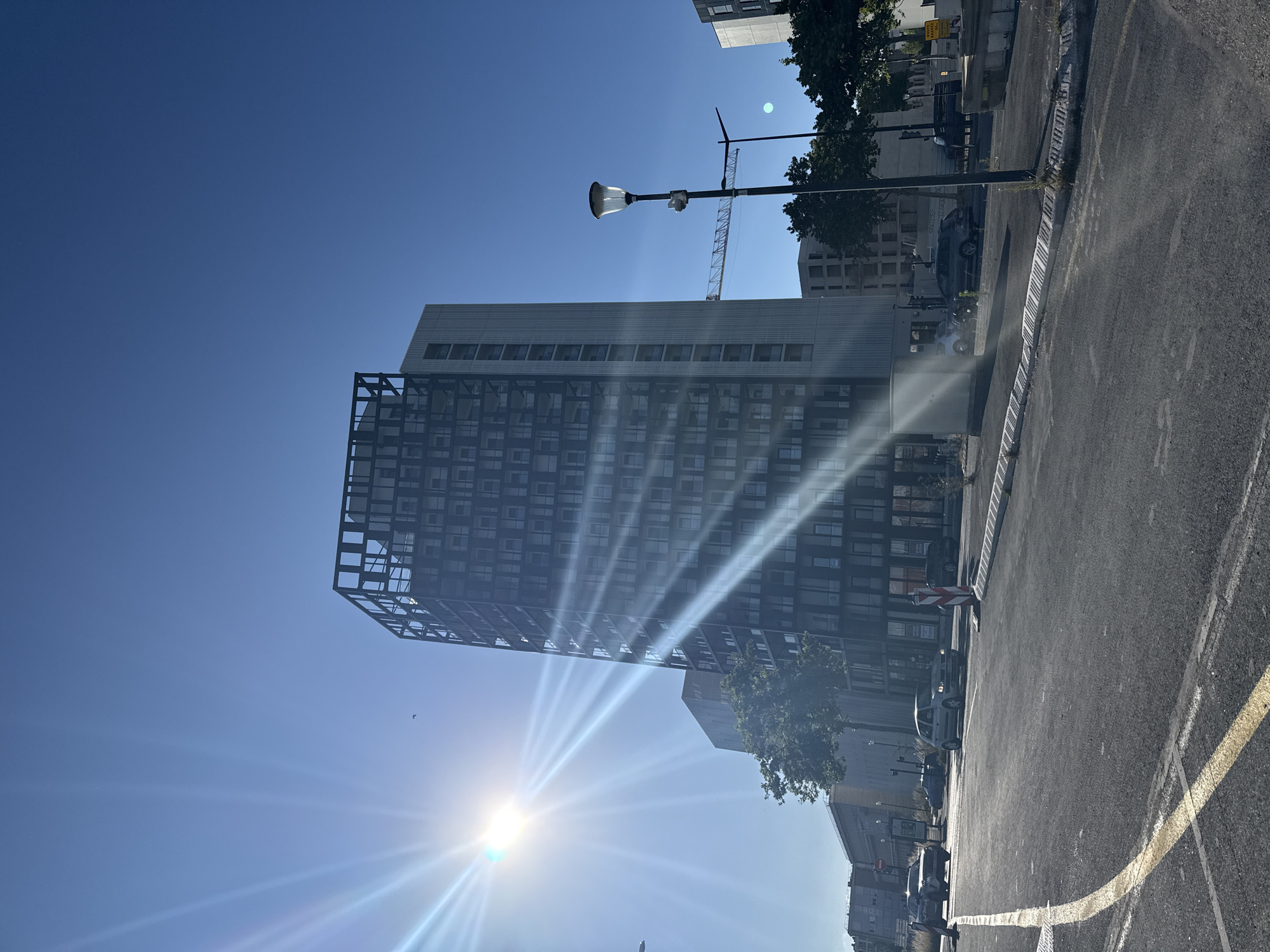 The image depicts a bright, sunny day in Le Havre, France, likely in a developing urban area. The foreground shows a newly paved road with yellow lane markings. Several cars are visible on the road, including a silver hatchback and a dark SUV. A tall, modern building with a dark facade and a skeletal structure on top dominates the midground, with sunlight streaming down its side, creating lens flare. To the right of this building, a streetlamp stands tall. In the background, other buildings, some appearing to be under construction, are visible, along with trees and a construction crane. The sky is a clear, vibrant blue.