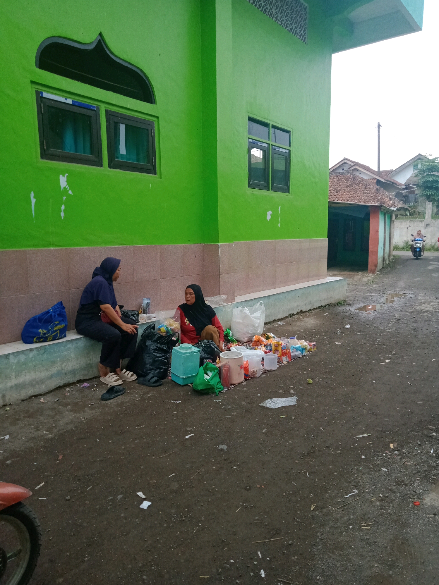 Two women are sitting on a concrete ledge in front of a bright green building with a patterned ventilation grate above two windows. The woman on the left is wearing a dark blue hijab and shirt with dark pants, and is sitting with her legs crossed. The woman on the right is wearing a black hijab and a red shirt with dark pants, and is facing the woman on the left. In front of them on the ground, there are several items spread out, including plastic bags, food items, and containers. A large black garbage bag is between the two women. A blue tote bag is to the left of the woman on the left. The ground in front of the building is paved and appears wet in places, with some scattered debris. In the background, a street with houses and a motorbike can be seen. This scene is located in Cilampunghilir, Indonesia.