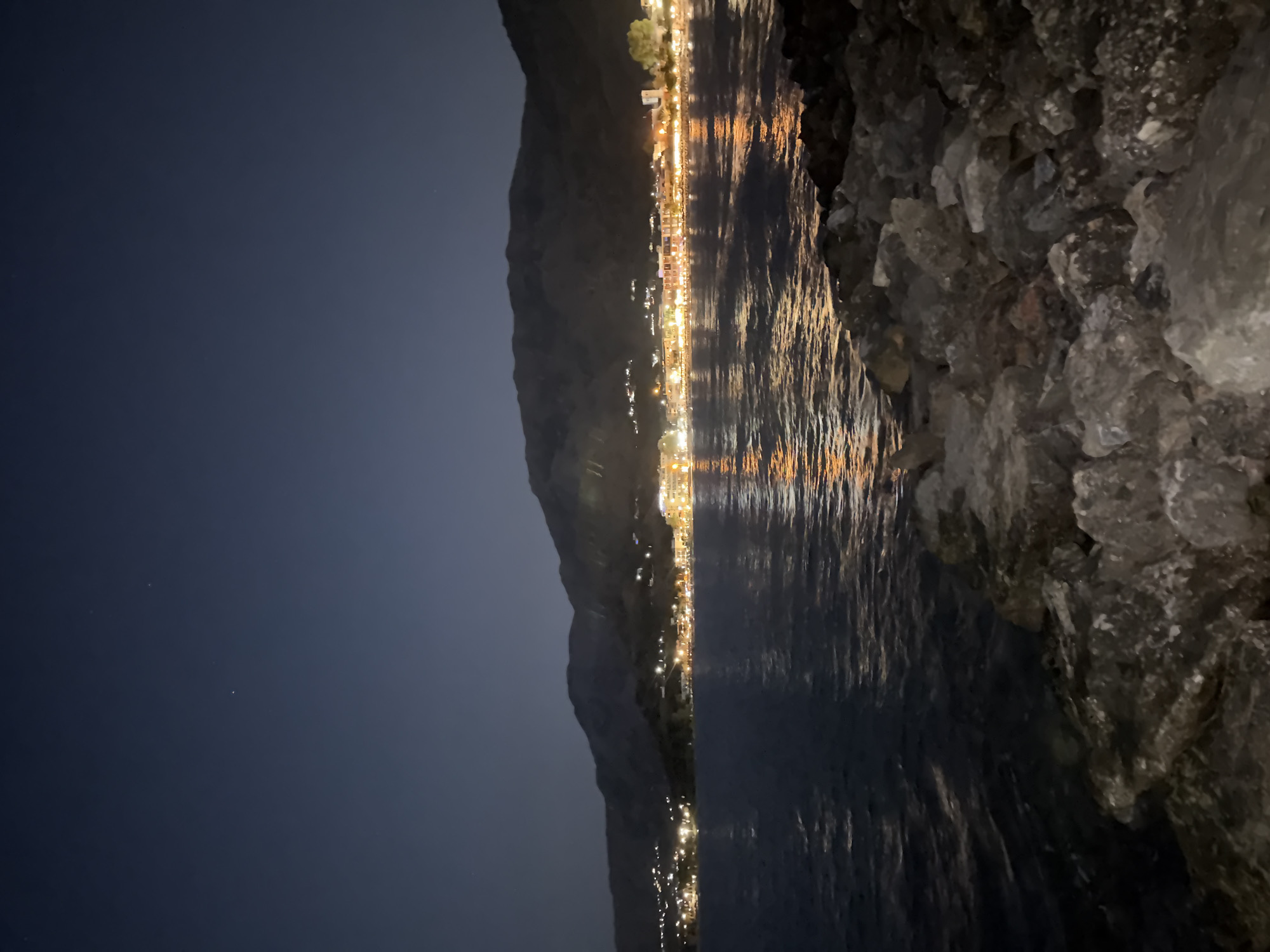 The image depicts a nighttime view of the town of Georgioupoli, Greece, from the water's edge. A rocky shoreline occupies the foreground, with dark, irregularly shaped stones leading down to the calm, dark water. In the midground, the sea reflects the warm, golden lights of the town that stretches along the coastline. The town is nestled at the base of a dark, undulating mountain range that rises in the background. Numerous lights indicate buildings and structures, suggesting a populated area. The sky above is a deep, dark blue, with a few faint stars visible. The overall impression is one of a tranquil coastal town illuminated by artificial lights under a clear night sky.