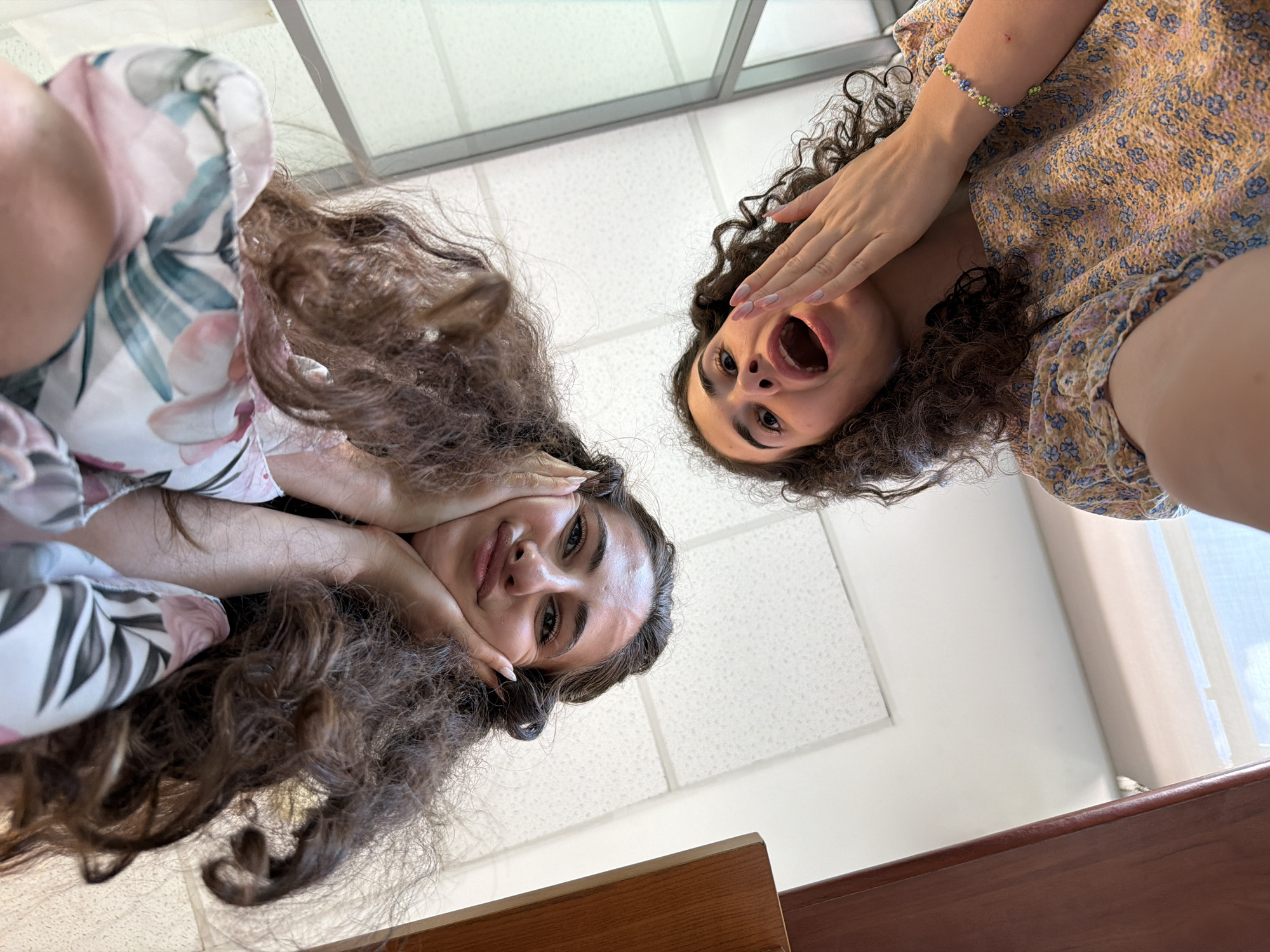 This image is a low-angle shot of two young women indoors. The woman on the left is wearing a white top with a floral pattern in pink and green. She has long, dark curly hair and her hands are touching her face. The woman on the right is wearing a light-colored, floral-patterned dress and has dark curly hair. Her right hand is covering her mouth, and her mouth is open in a surprised expression. A beaded bracelet is visible on her left wrist. The background appears to be an indoor ceiling with textured tiles and fluorescent lighting. The foreground shows a wooden surface, likely a table or desk. The perspective suggests the photo was taken from below, looking up at the subjects. No specific city or country is identifiable from the visual cues in the image.