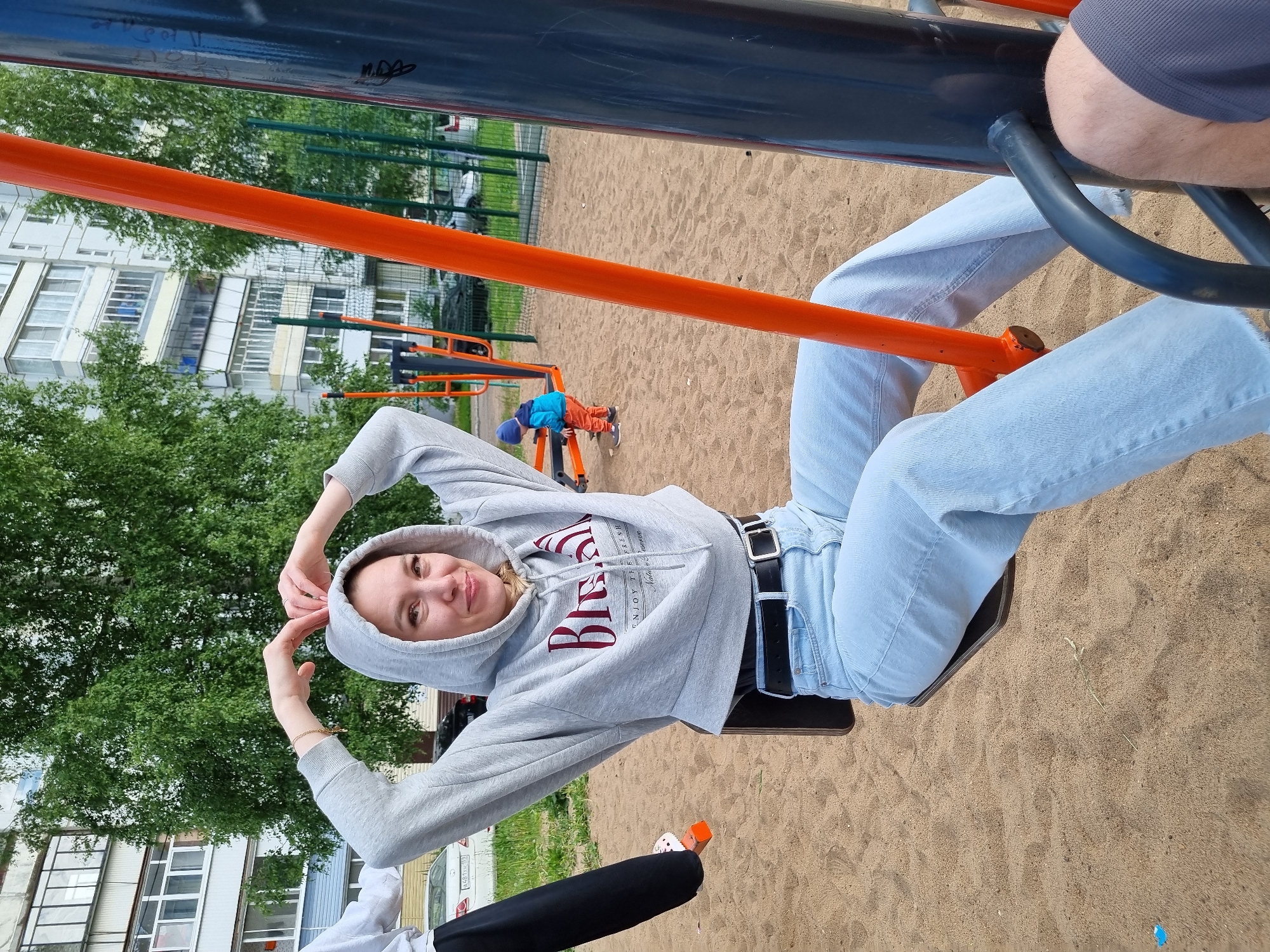 An eye-level medium shot captures a woman sitting on a swing at a playground. She is wearing a grey hooded sweatshirt with red lettering, light blue wide-leg jeans, and a black belt. Her hands are raised, forming a heart shape above her head, and she is smiling at the camera. In the background, a young child in a blue jacket and orange pants plays on playground equipment. Beyond the playground, apartment buildings and trees are visible. The scene is set in a sandy playground area, likely within the city of Chaikovsky, Russia, given the architecture and general environment. The lighting suggests an overcast day.