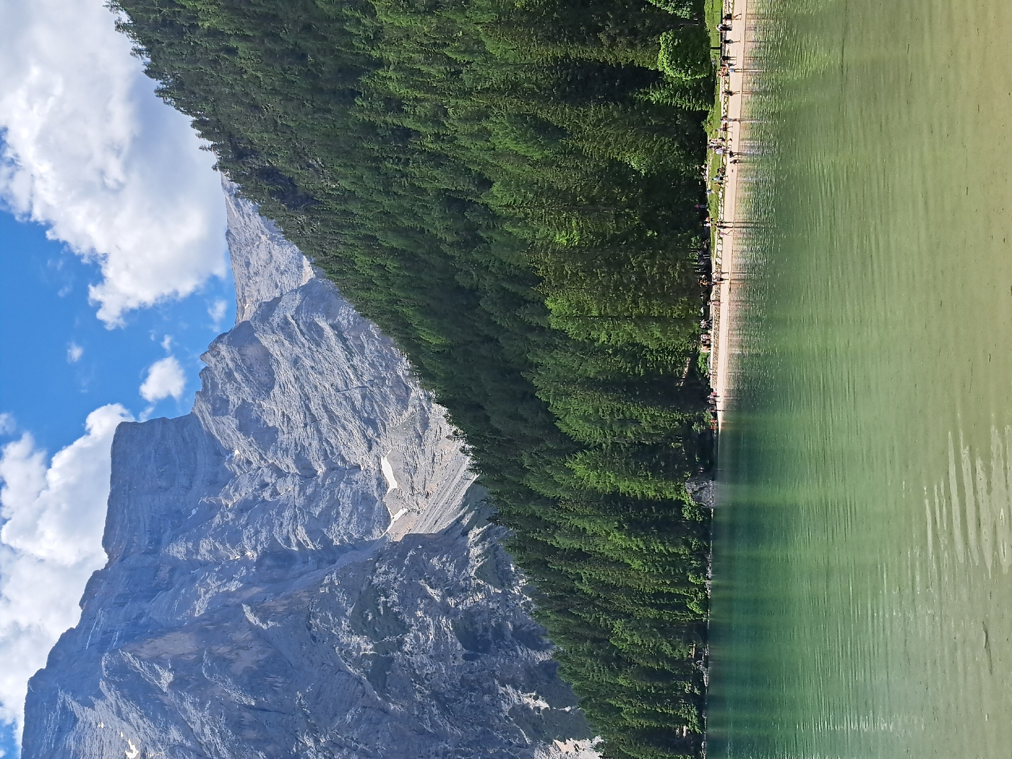 This is a landscape photograph taken from a high vantage point, looking down at a tranquil lake with a forested mountain backdrop. The image is oriented vertically, with the lake and its shoreline occupying the right side and the mountains and trees on the left.
The lake is a clear, pale green, with subtle ripples on its surface indicating calm conditions. Along the shoreline, a sandy path is visible, lined with numerous people walking and standing, enjoying the scenery. They appear as small figures from this height, some casting long shadows that stretch towards the water, suggesting it is likely late afternoon or early morning. A dense, dark green forest of pine trees forms a thick wall behind the path, climbing up the lower slopes of the imposing mountain range. The mountains themselves are rugged and grey, with steep, rocky faces and patches of snow or scree on their upper reaches. The sky above is a bright blue, dotted with fluffy white clouds, indicating pleasant weather.
The scene evokes a sense of peacefulness and natural beauty, with the combination of the serene lake, lush forest, and majestic mountains creating a picturesque setting. The presence of people suggests a popular tourist destination or recreational area. No text is visible in the image.