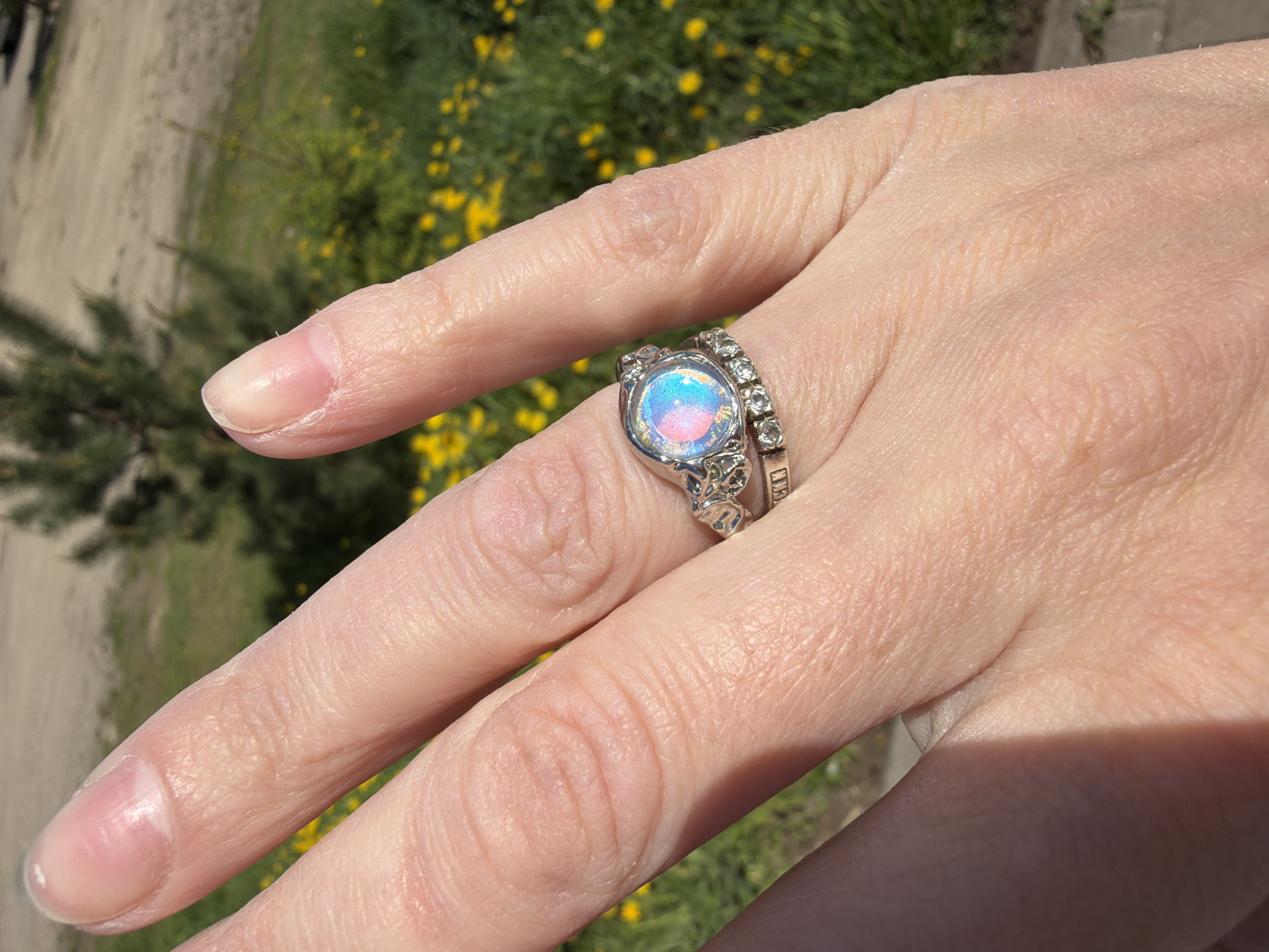 A human hand is positioned in the foreground, adorned with two rings on the ring finger. The primary ring features a round, iridescent gemstone encircled by a silver-colored setting. Adjacent to it is a thinner band of small, clear stones set in a matching silver-colored metal. The fingers are naturally shaped with well-manicured, oval-shaped nails. The skin appears to be light-toned and finely textured. The background is an out-of-focus outdoor setting, with greenery and yellow flowers. The lighting suggests a sunny day. The location is not specified in the provided image information.