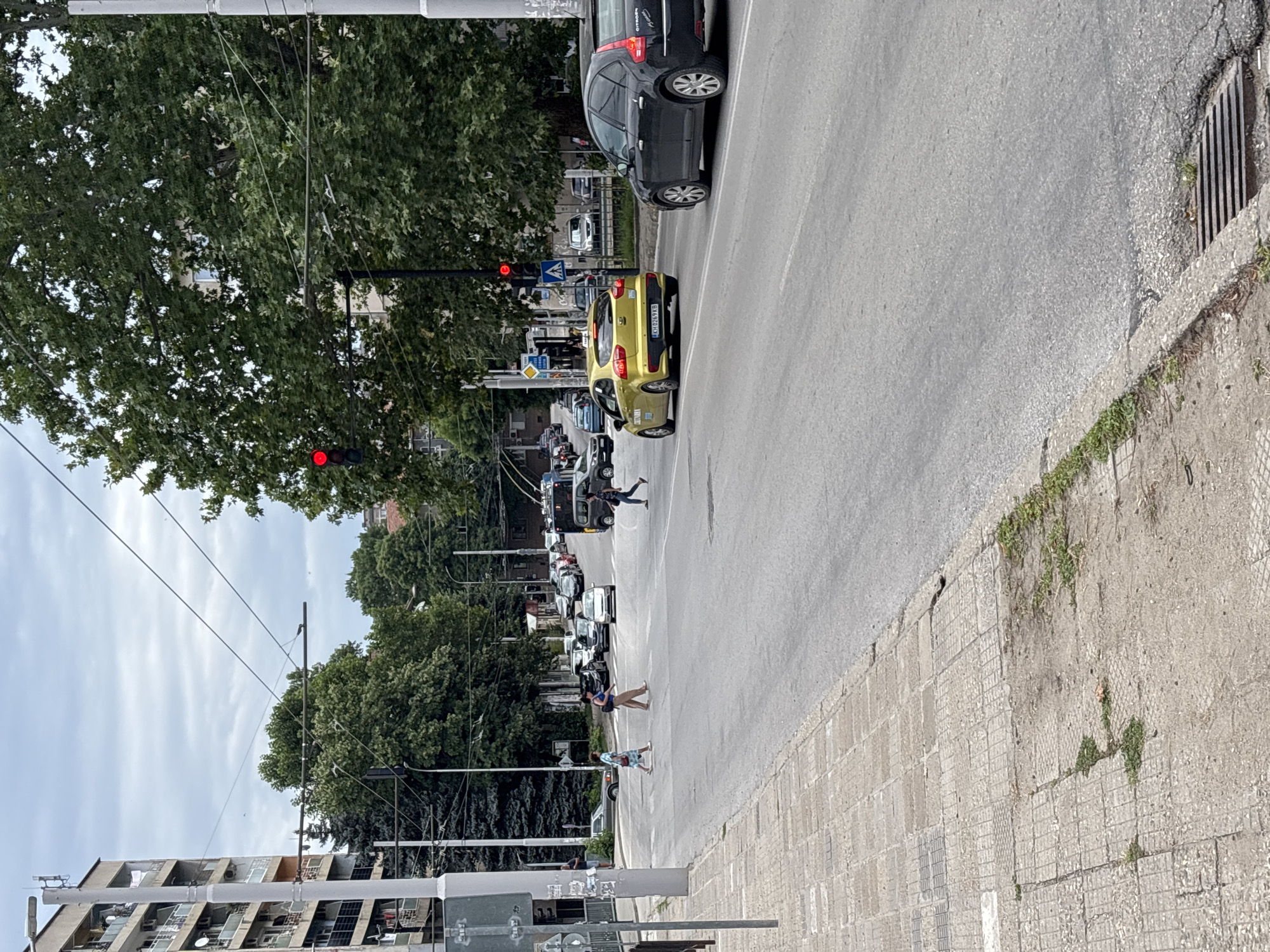 The image depicts a street scene in Pleven, Bulgaria, likely on a cloudy day. In the foreground, a paved sidewalk with scattered patches of grass runs along the left side of a multi-lane asphalt road. Several pedestrians are visible crossing the road at a designated crosswalk.
Dominating the mid-ground are several vehicles. A yellow Kia Rio taxi with license plate "KH 8269 KB" is driving on the right lane of the road. To its right, a dark-colored Citroën hatchback is also in motion. Further down the road, a line of cars and a dark-colored van are stopped, presumably at a traffic light.
In the background, apartment buildings and lush green trees line the sides of the street. Overhead, power lines and what appear to be trolleybus wires are suspended across the road. A traffic light displaying a red signal is visible above the yellow taxi, indicating that vehicles should stop. The overall environment suggests a typical urban setting within the city.