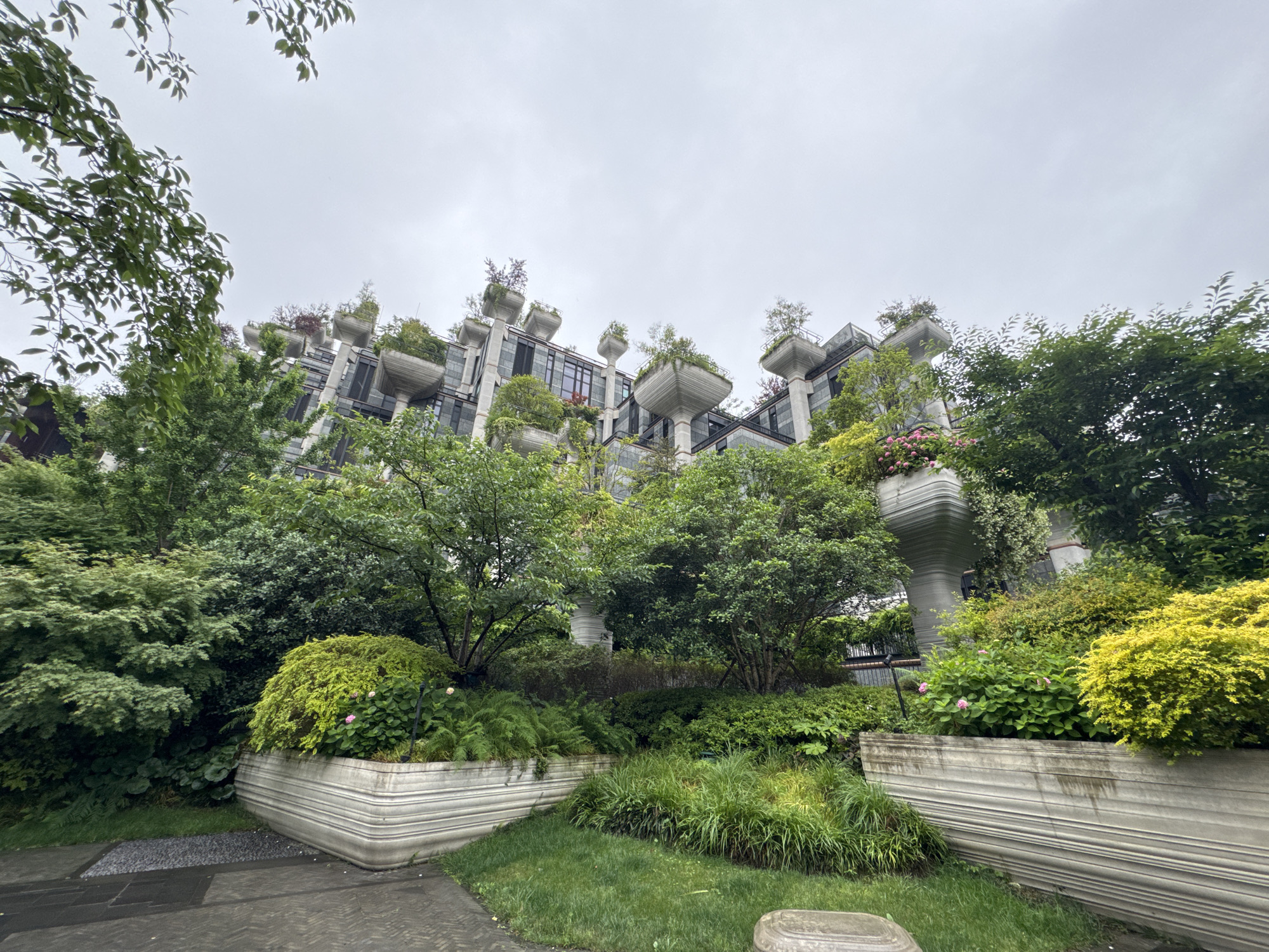 The image depicts a building in the Putuo District of Shanghai, China, distinguished by its unique architecture and extensive greenery. The building has a contemporary design with gray stone, glass windows, and individual balconies or terraces supported by fluted, pillar-like structures. Each balcony has a large planter filled with trees, shrubs, and flowering plants, creating a "hanging gardens" effect. In the foreground, there's a well-maintained garden with stone planters filled with a variety of plants, bushes, and trees. The lawn is manicured, and there are stone pathways visible. The sky is overcast, suggesting a cloudy day.