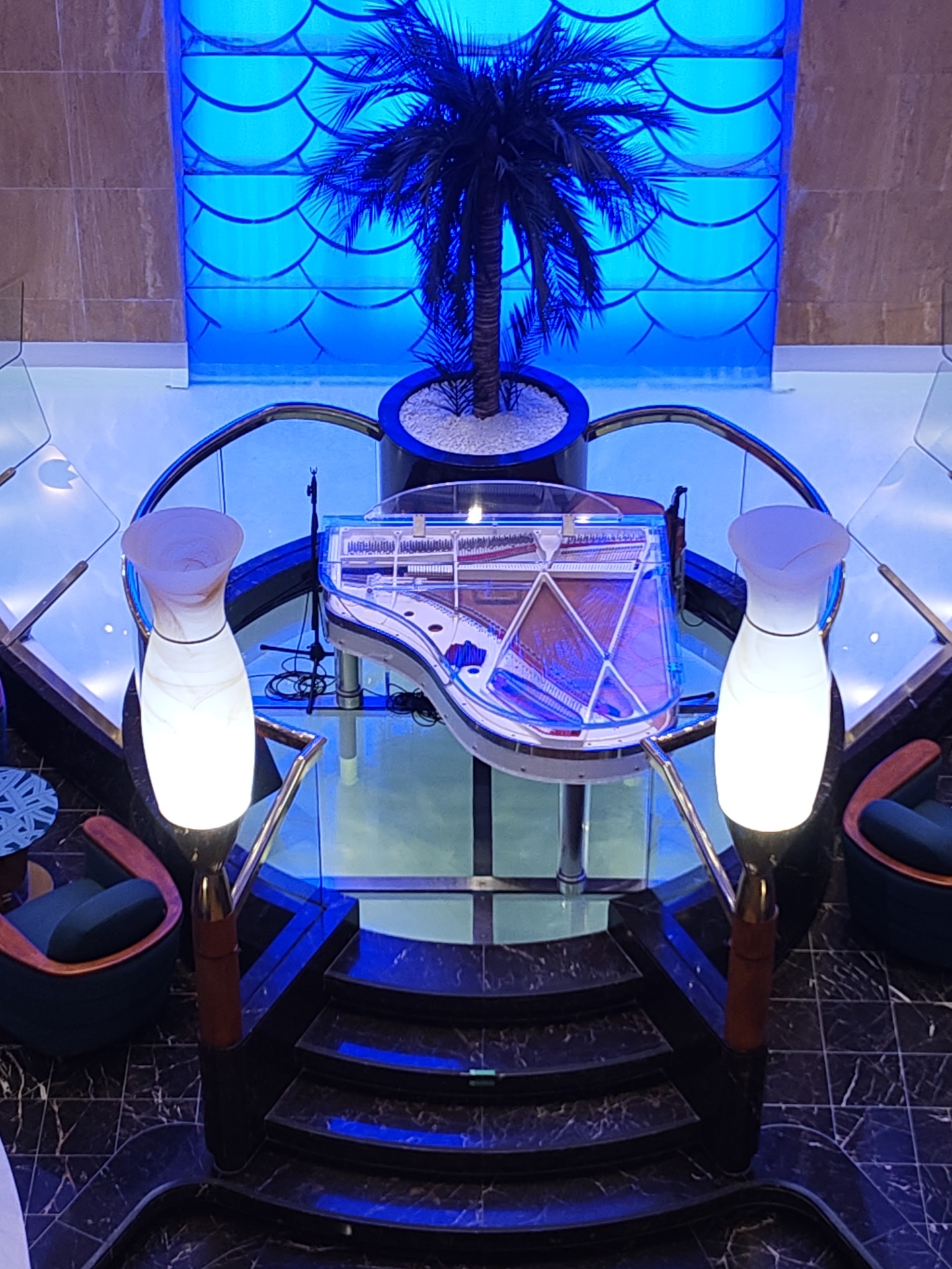 Captured from an elevated perspective, the image showcases an ornate interior scene in the city of Unknown, Unknown. At the center is a transparent grand piano, encircled by a rounded railing and situated above a series of dark-colored steps. Adjacent to the steps are two standing lamps with white, vase-shaped shades. Two plush chairs with wooden frames are placed on either side of the piano setup. A potted palm tree is situated in the background, set against a wall with a unique scale-like pattern and bright blue lighting. The floors are a polished, marble-like material.