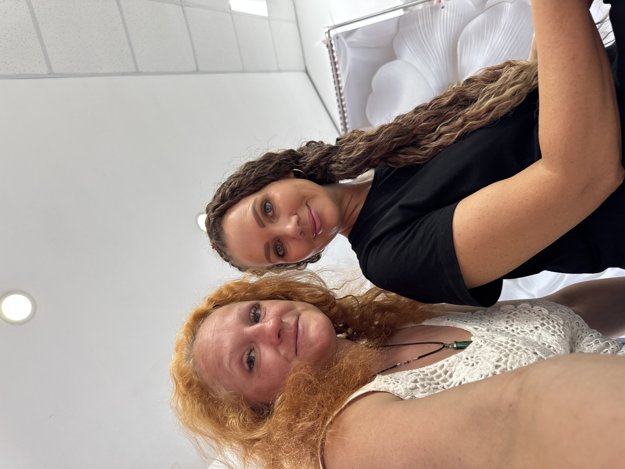 Two women are posing for a selfie indoors. The woman on the left has wavy, ginger-colored hair and is wearing a white, crochet-like top with a necklace featuring a green pendant. The woman on the right has long, braided, brown hair and is wearing a black t-shirt. Behind them, a white, decorative object, possibly the interior of a coffin or a large floral arrangement, is visible. The ceiling appears to be a suspended grid system, and there is a circular light fixture. The image quality is adequate, and the lighting is bright. The scene does not provide any visual cues to determine a specific location within the city of Chaikovsky, Russia.