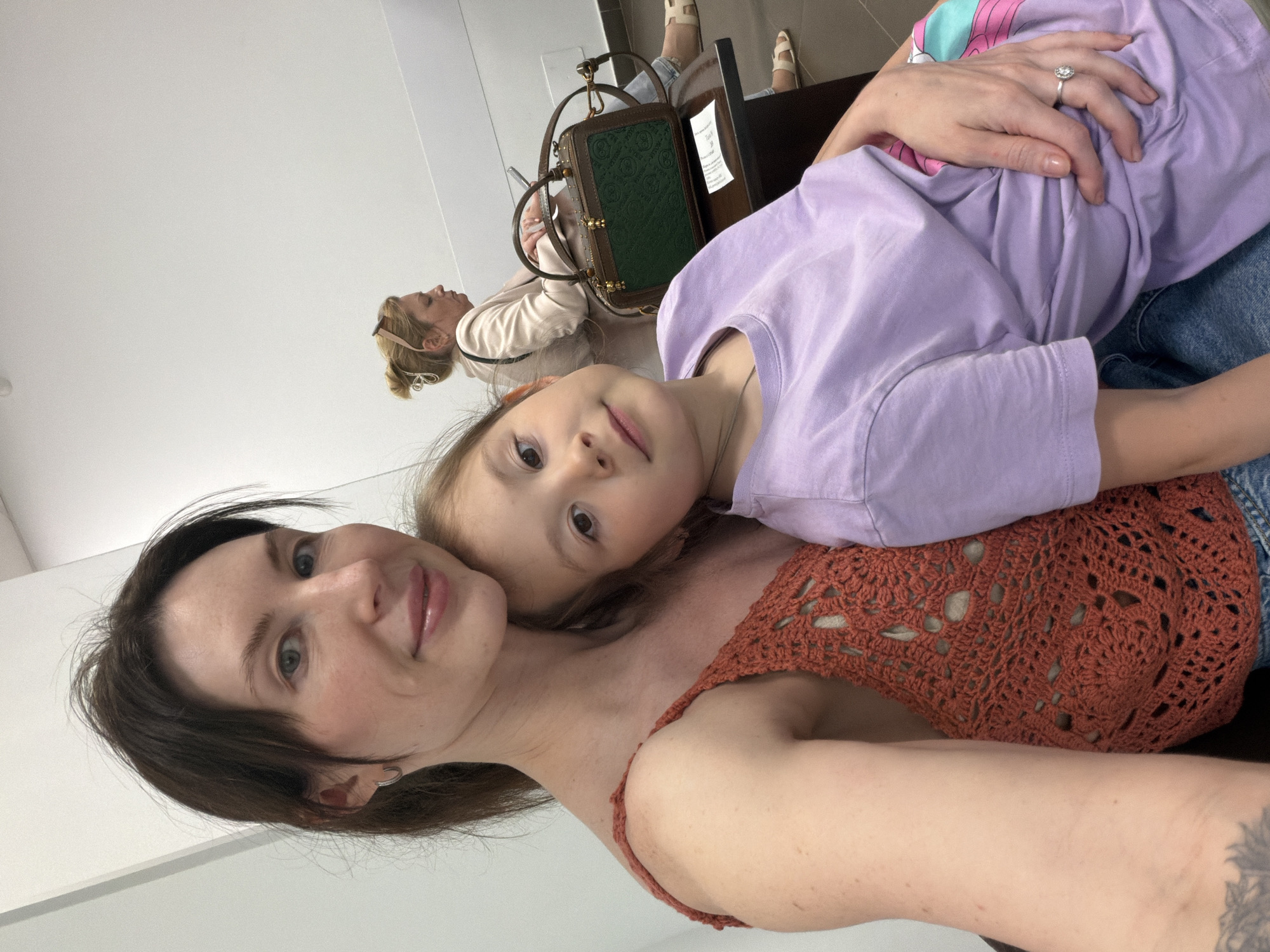 The image shows a selfie of a woman and a young child. The woman has dark hair, fair skin, and is wearing an orange crocheted top. The child is lying on the woman, facing the camera, and wearing a purple t-shirt. Behind them, another woman is sitting at a table with a purse. The location is in Perm, Russia.