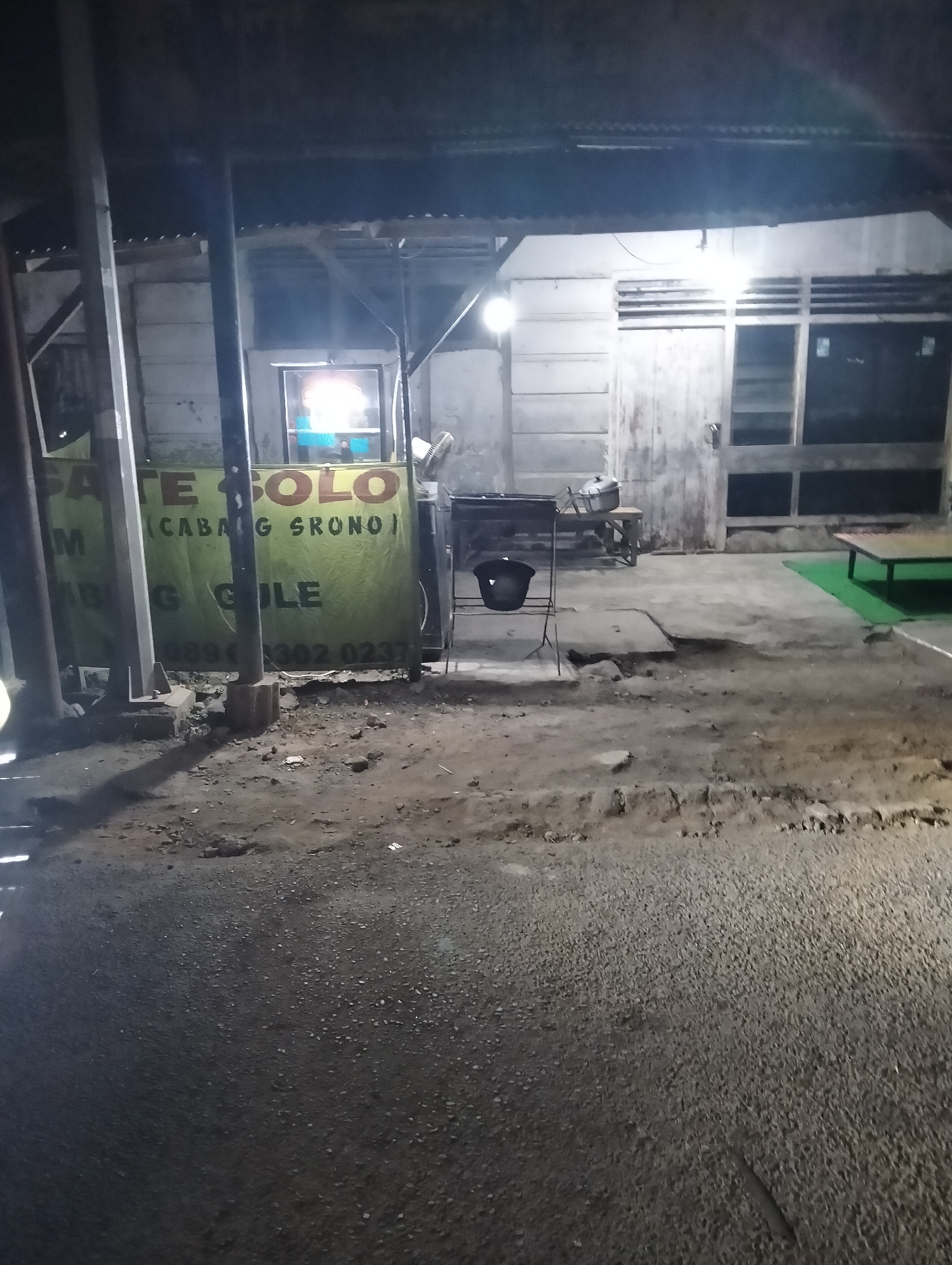 A photograph taken at night depicts an outdoor food stall or eatery in Blambangan, Indonesia. The stall features a green banner with text in Indonesian, including "Sate Solo," "(Cabang Srono)," and "Gule." The setting appears to be a dimly lit street or outdoor area with a concrete ground and some patches of dirt and gravel. In the background, there is a building with horizontal-slatted windows and a wooden door. A metal grill and a black bucket are visible near the stall. A small green mat and a low table are also present in the scene. The overall impression is of a modest, local establishment operating in the evening.