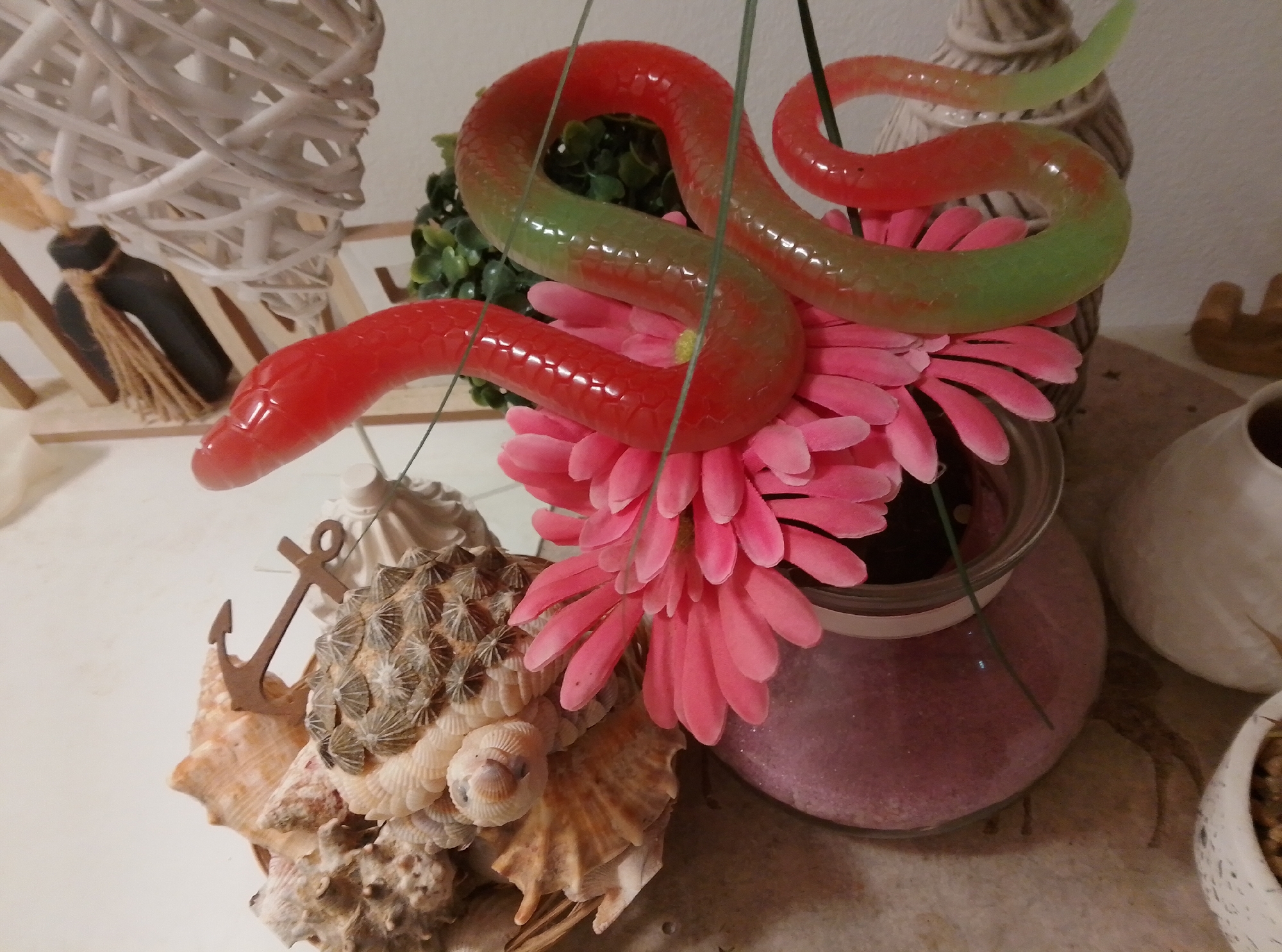 A diorama is displayed on a white surface. In the center of the diorama, a red and green translucent snake sculpture is suspended by thin wires, appearing to coil around a large pink artificial flower. The snake's body transitions from red at the head to green towards its tail. The pink flower rests on a clear glass bowl filled with pink decorative sand. To the left of the flower, a collection of seashells and barnacles are arranged, with a small wooden anchor figurine placed on top of them. In the background, to the left, a white woven basket structure is visible, with a dark object and twine hanging from it. To the right of the snake, a beige, textured vase is partially visible, and further to the right, a white, rounded ceramic object and a small white pot with soil are present. The overall scene suggests a decorative arrangement, possibly for display or as part of a themed setting. This scene is not located in the city of Wedel, Germany; it is an image of decorative objects on a white table.