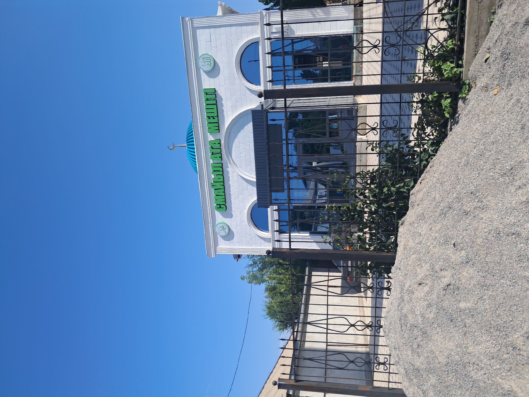 The image shows the Sayat Ata mosque in Shymkent, Kazakhstan, on a clear, sunny day. The mosque is a modern white building with a blue dome topped by a crescent moon and star. Green Arabic script on the facade reads "Sayat Ata Meshiti". The mosque is enclosed by a black metal fence with ornate scrollwork. In front of the fence, a pile of gravel is visible. A car is parked behind the fence. Green trees and vegetation are also present in the scene. The sky is a vivid blue.