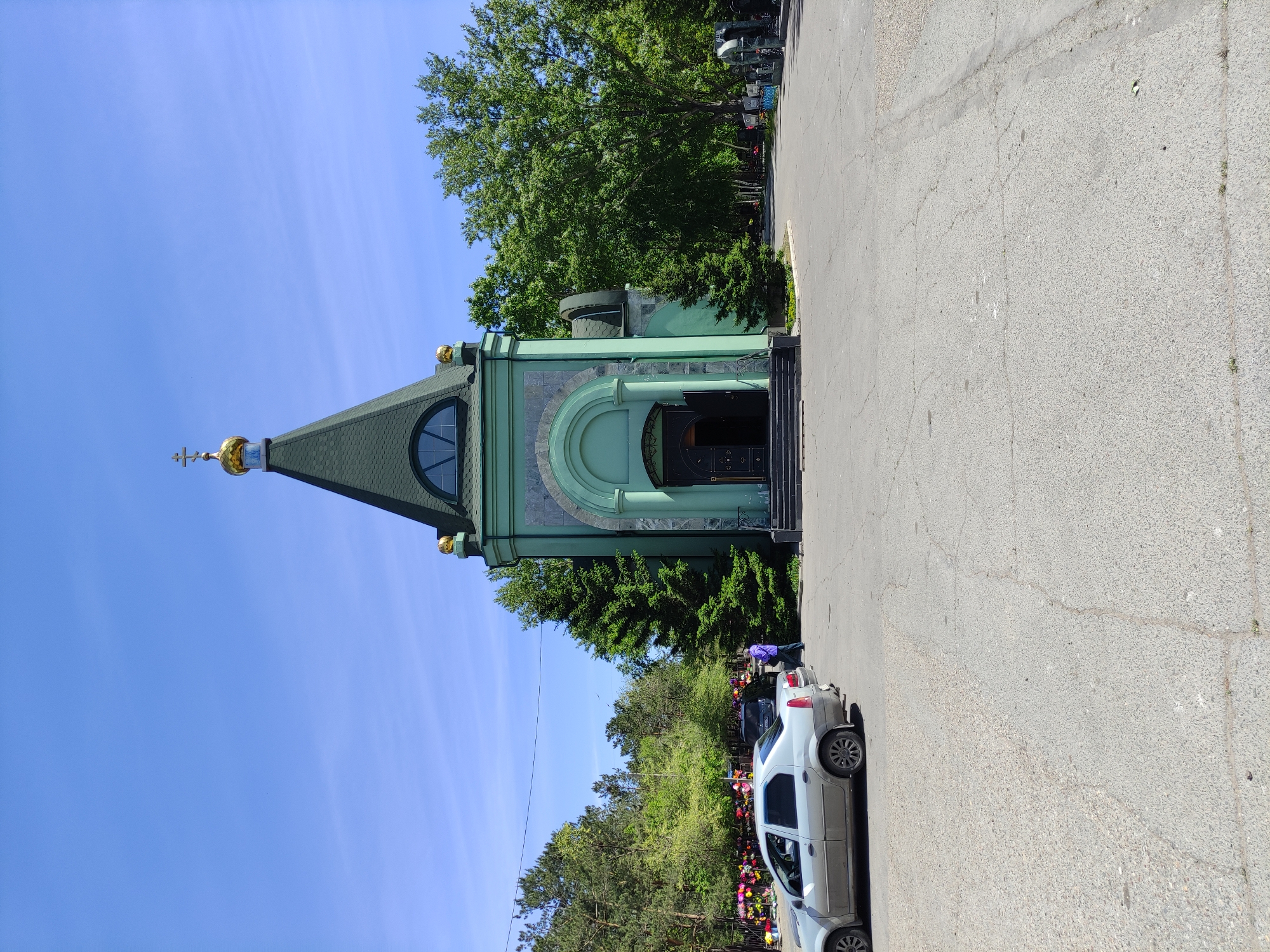 The image shows a light-green, ornate church structure with a dark-green, pointed roof and a gold cross on top. The church has a large, arched entrance with dark-colored steps leading up to it. To the right of the church is a wide, gray, paved road with cracks. A white car is parked along the edge of the road, partially obscuring a view of what appears to be a cemetery with numerous flower decorations behind it. Lush green trees surround the church and cemetery area. The sky is clear blue with faint streaks. It is located in Pavlodar, Kazakhstan. A person in purple is standing near the car.