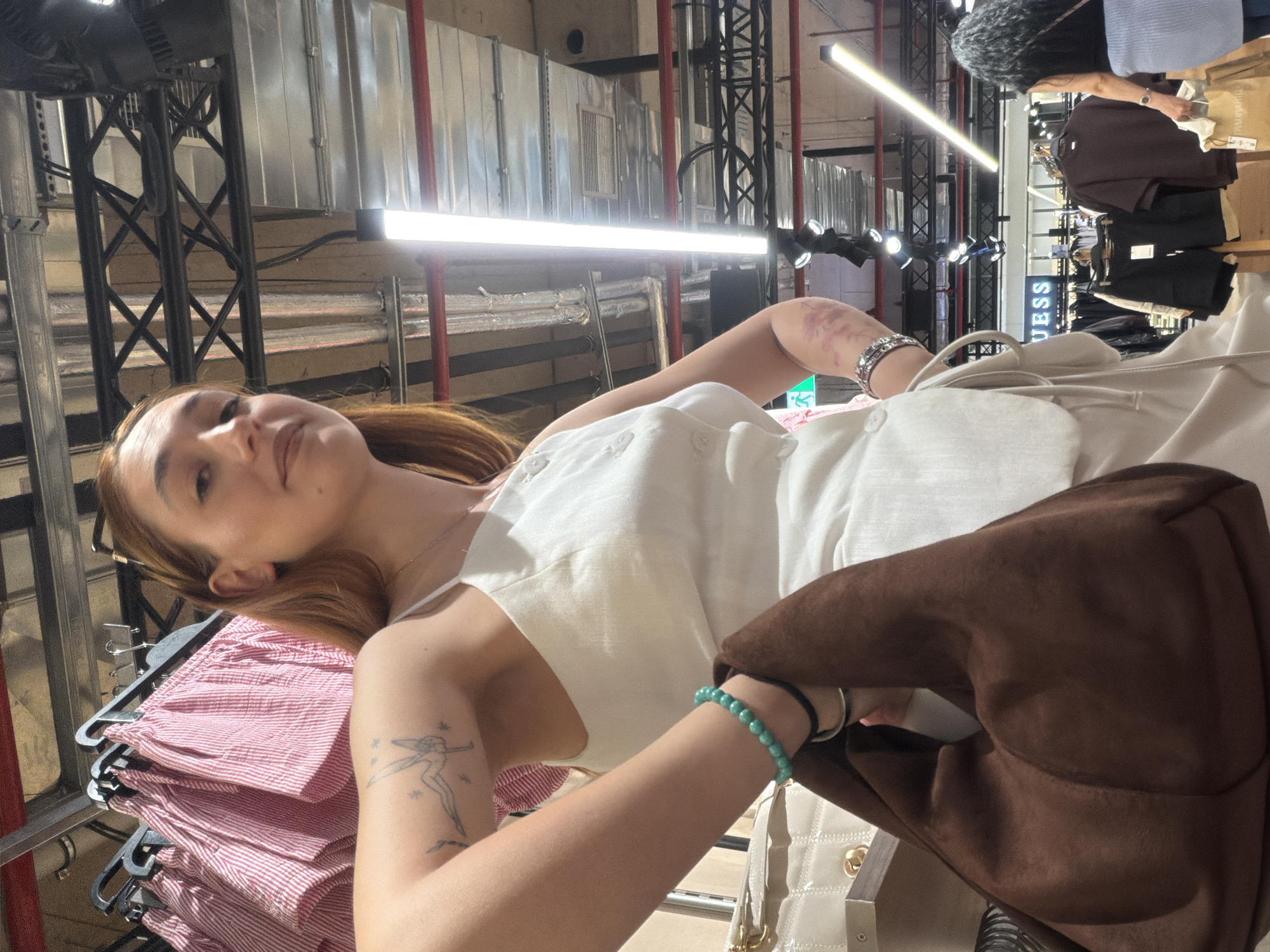 A woman is in what appears to be a clothing store, evident from the racks of clothes surrounding her. She is wearing a white dress or top and has a brown jacket or bag draped over her shoulder. There is a bracelet visible on her left wrist. Her arms feature tattoos. In the background, clothing racks and store fixtures, including a "GUESS" sign, are seen. The environment suggests an indoor retail space. I have no information to confirm this is in Sofia, Bulgaria.